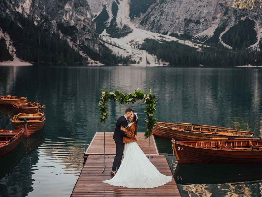 Bride and groom kissing under floral arch on wooden dock with mountain lake and rowboats backdrop.