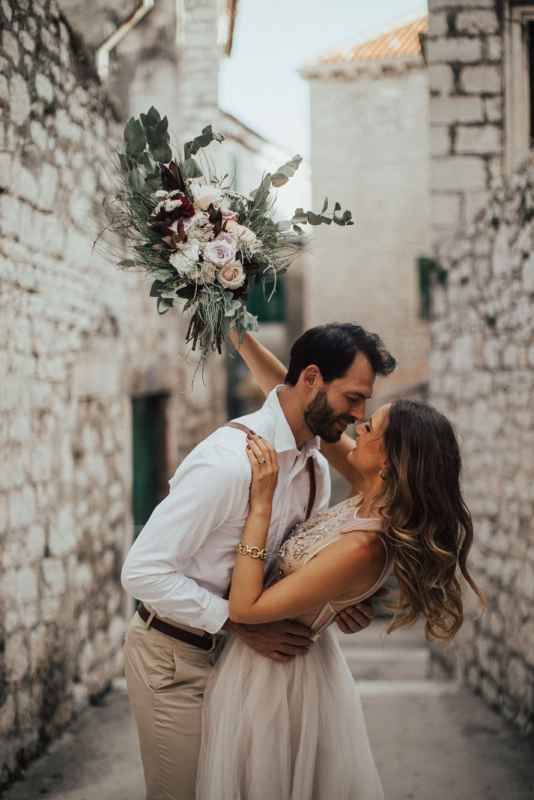 Couple shares a romantic moment in a rustic stone alley, bride holding a lush floral bouquet.