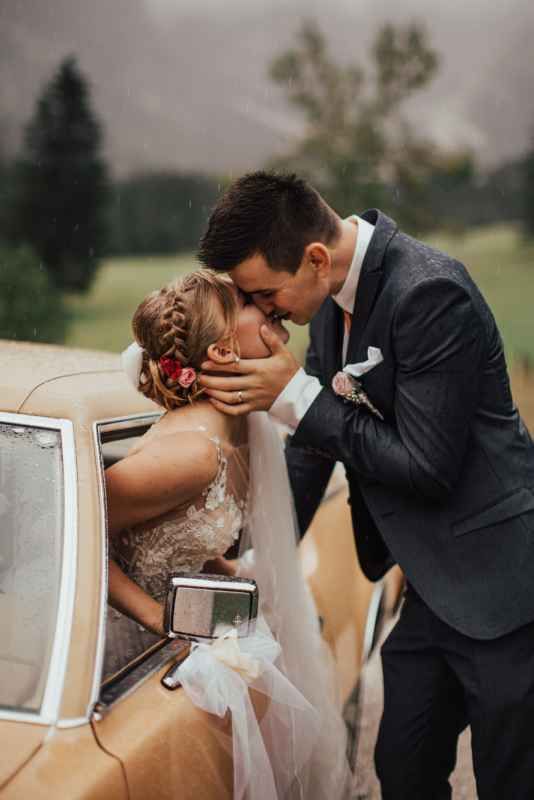 Bride and groom sharing a passionate kiss through a vintage car window in the rain.