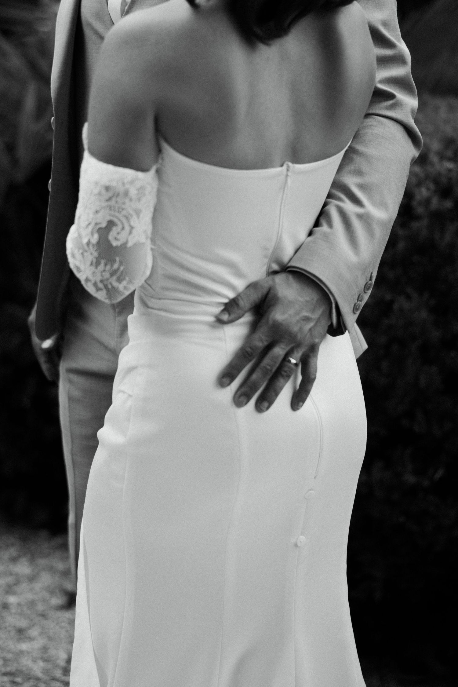 Black and white photo of bride in strapless gown with groom's hand on her back, wearing wedding ring.