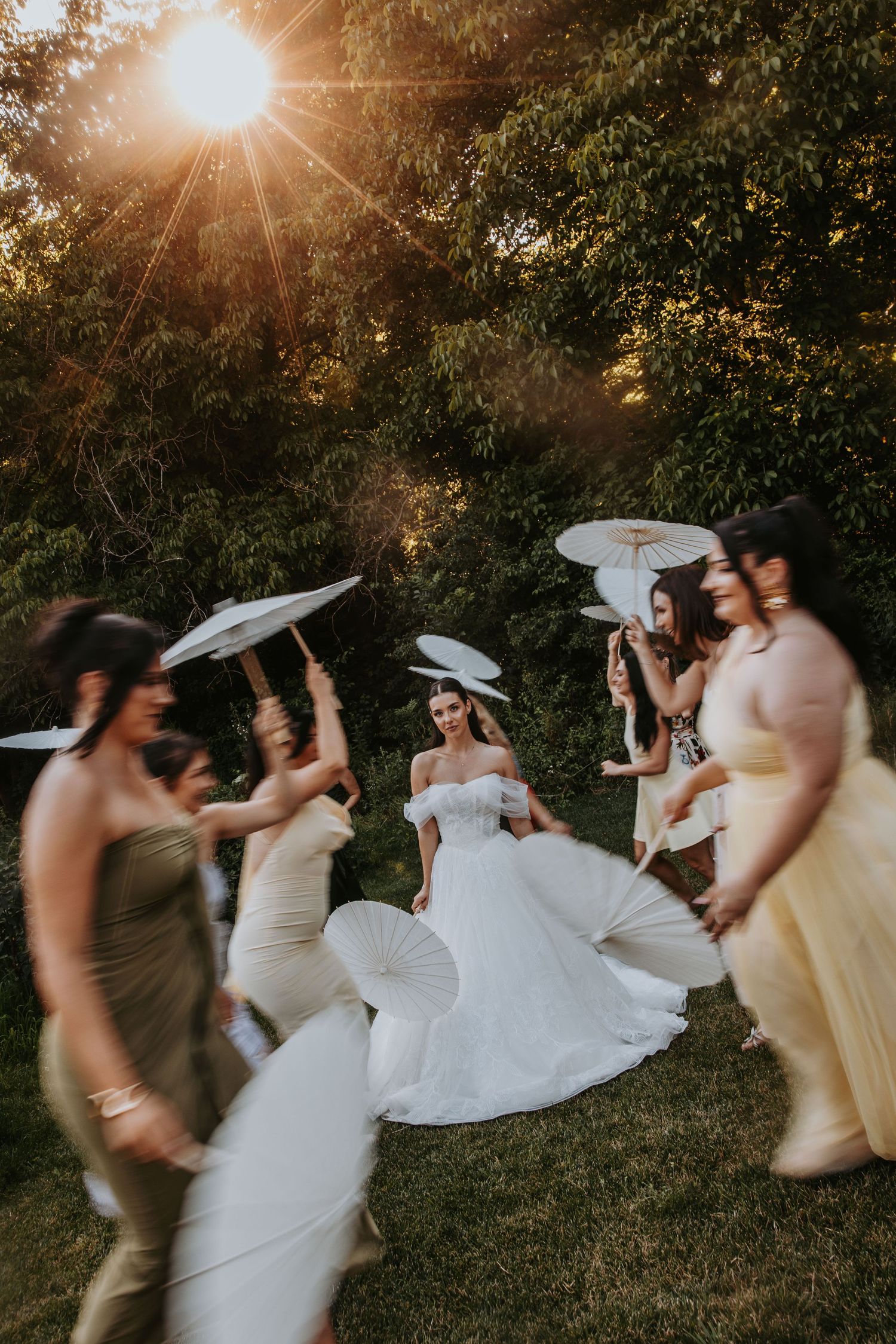 Bride in white gown surrounded by bridesmaids with parasols dancing in golden sunset light outdoors.