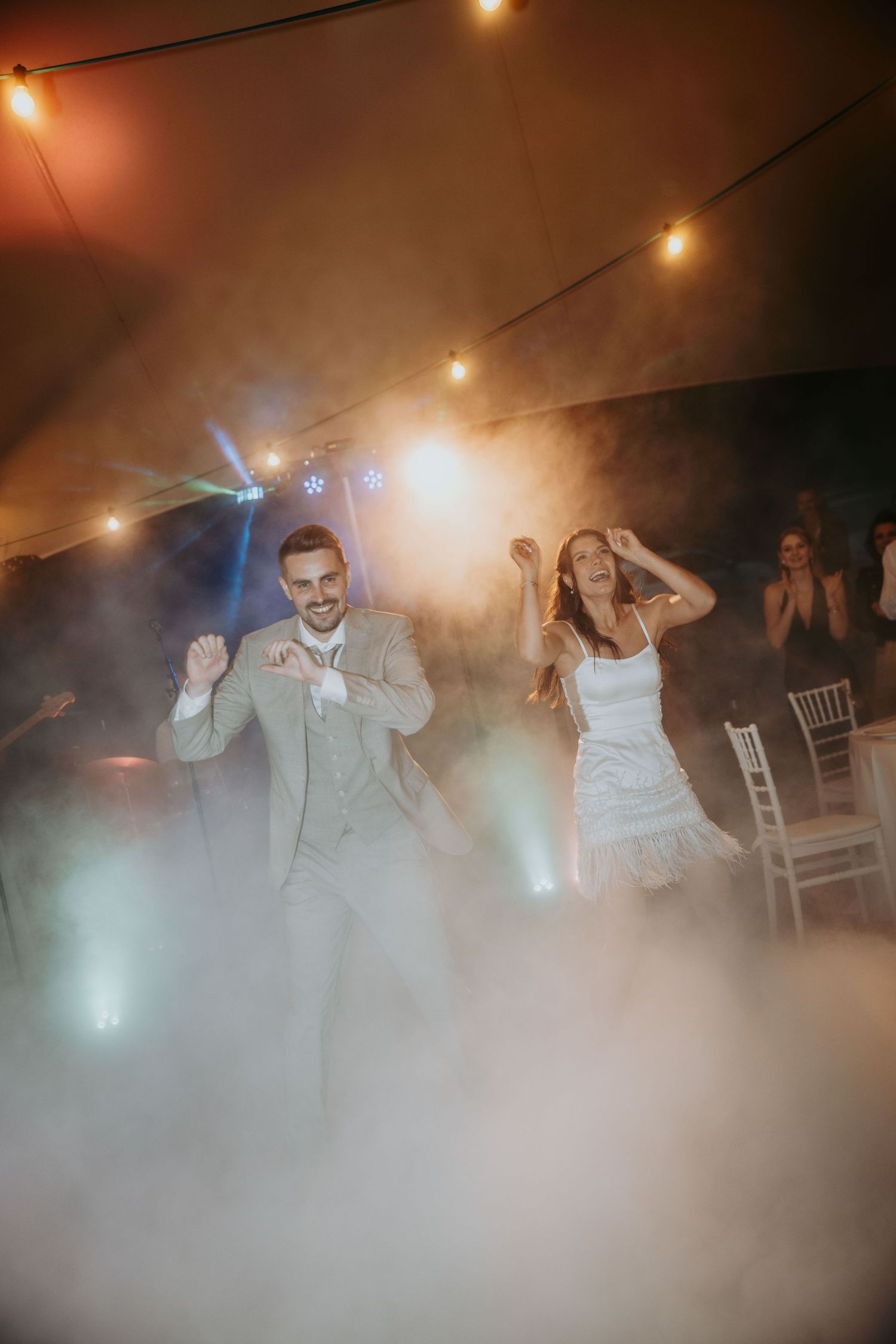 Bride and groom dance joyfully on a fog-filled dance floor under warm string lights at wedding reception.