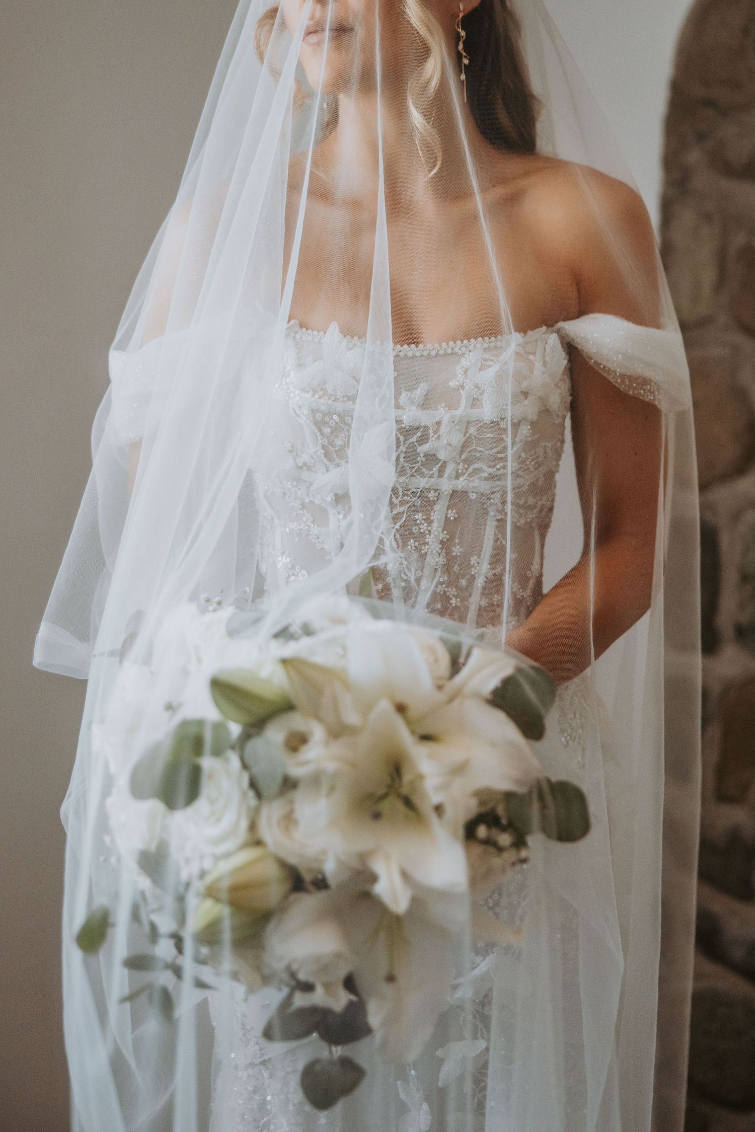 Bride in off-shoulder lace wedding gown holding white lily bouquet with veil draped over her face.