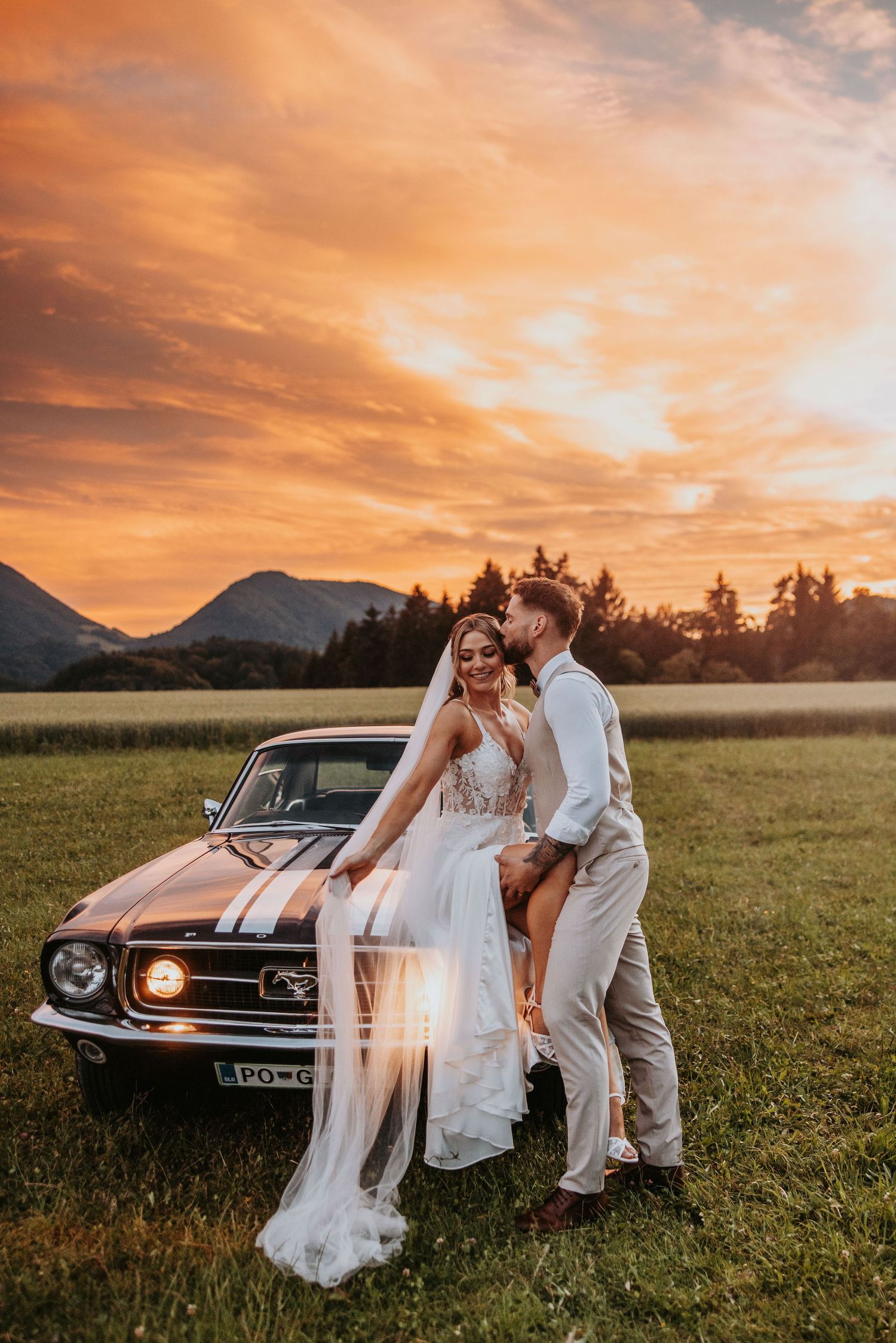 Bride and groom pose by vintage Ford Mustang at sunset with mountains in background.