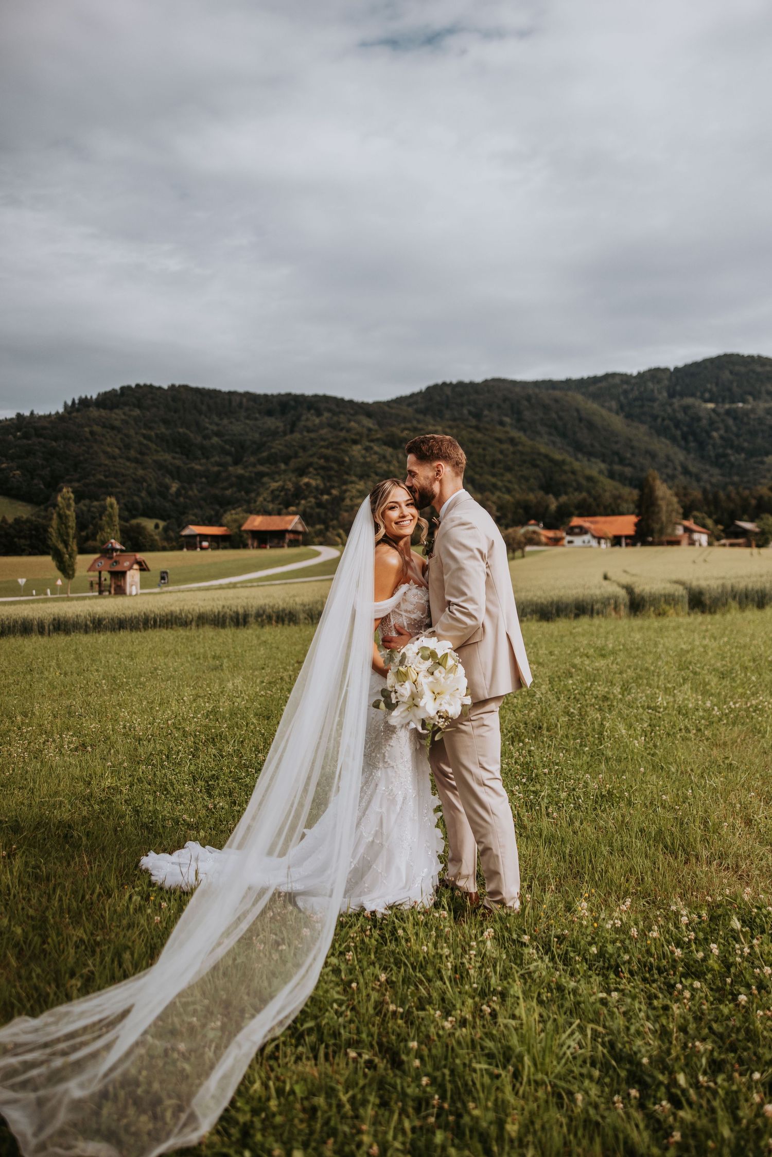 Bride and groom embracing in green meadow with mountain backdrop, long veil trailing behind.