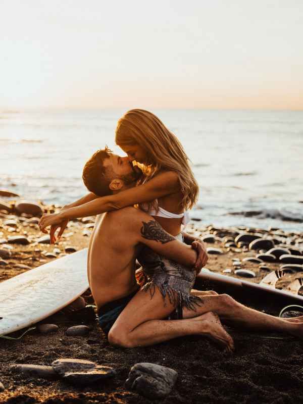 Couple sharing a romantic moment on a rocky beach at sunset with a surfboard nearby.