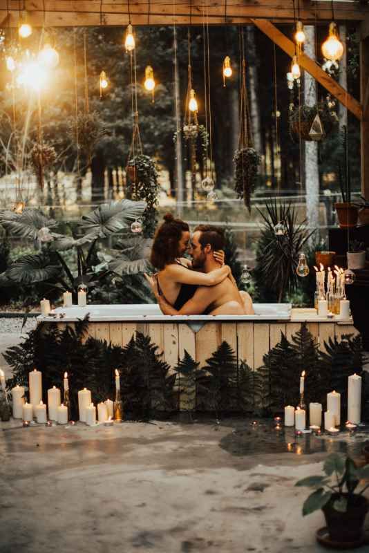 Romantic couple kissing in candlelit bath surrounded by lush plants and hanging Edison bulbs.