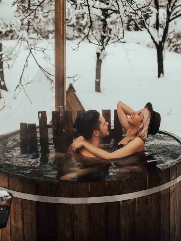 Romantic couple embracing in outdoor wooden hot tub surrounded by snowy winter landscape.