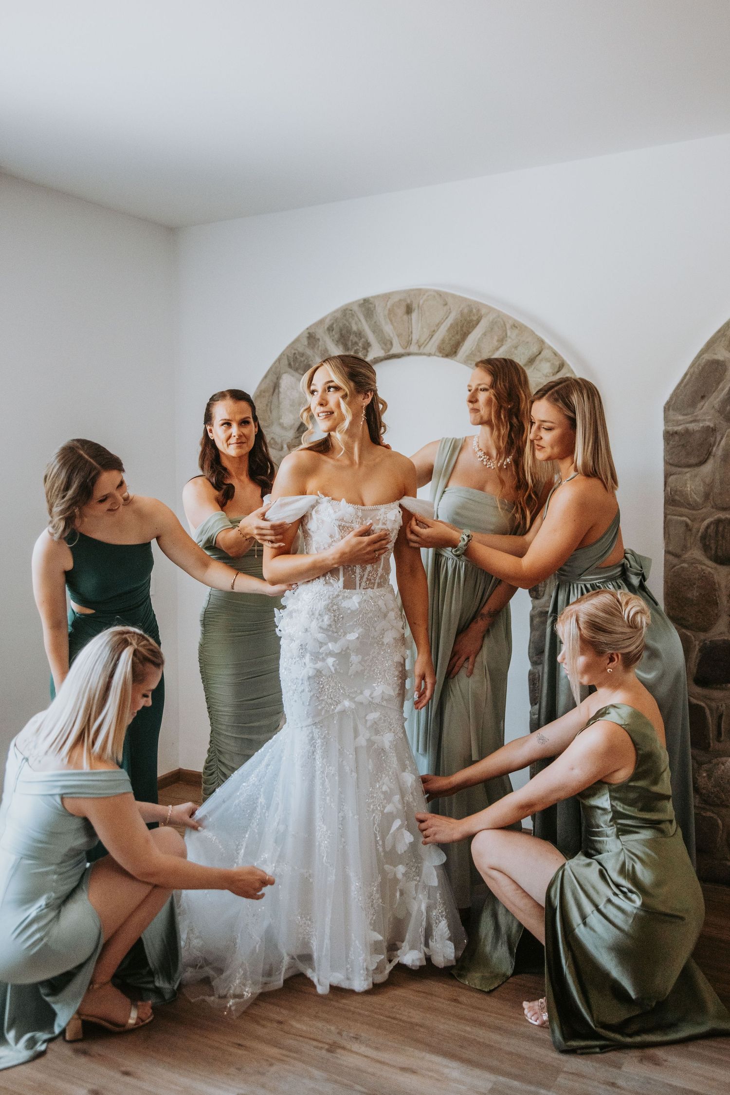 Bride in floral lace gown surrounded by five bridesmaids in green dresses helping her get ready.