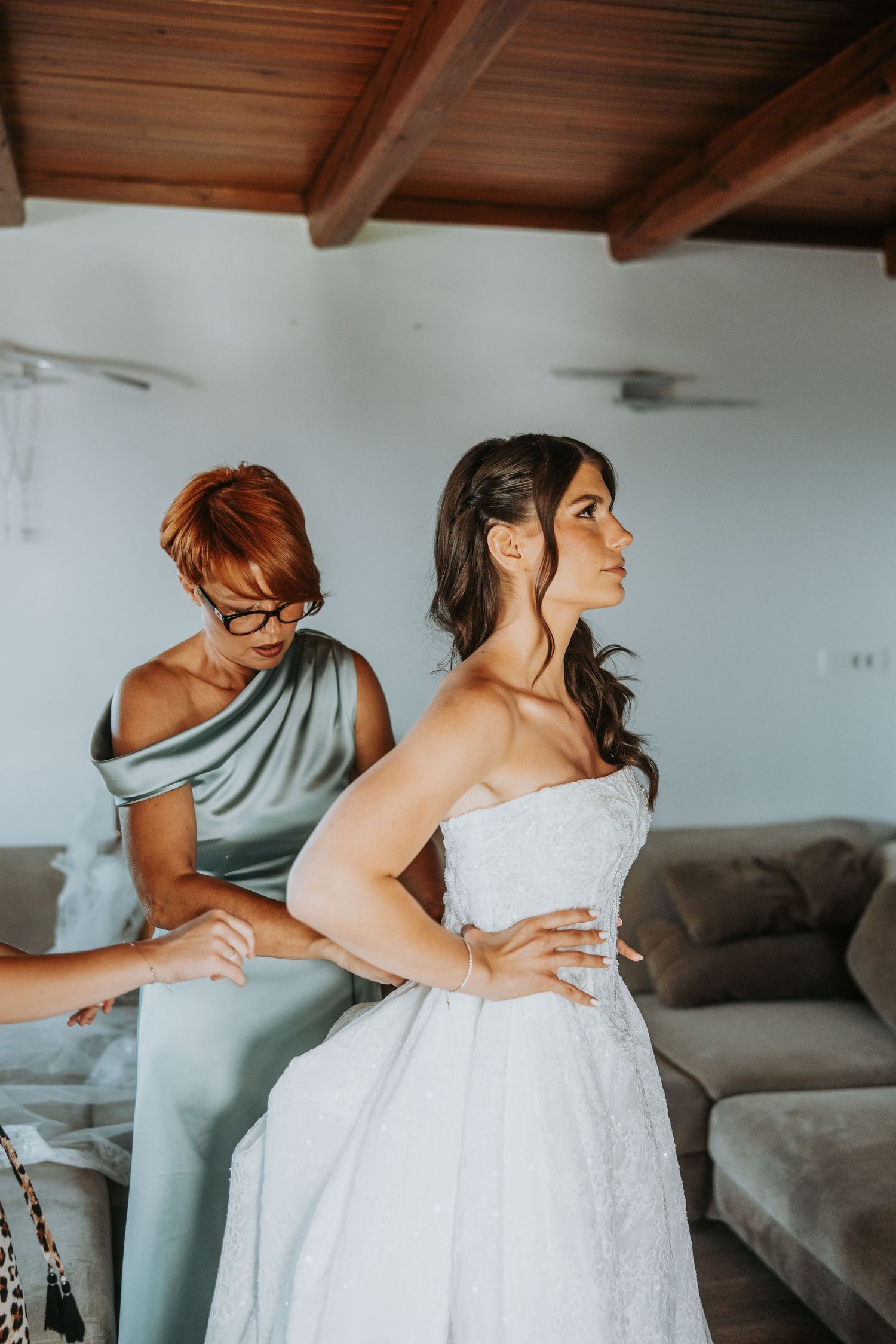 Bride in white lace gown gets help dressing from a woman in sage green dress indoors.