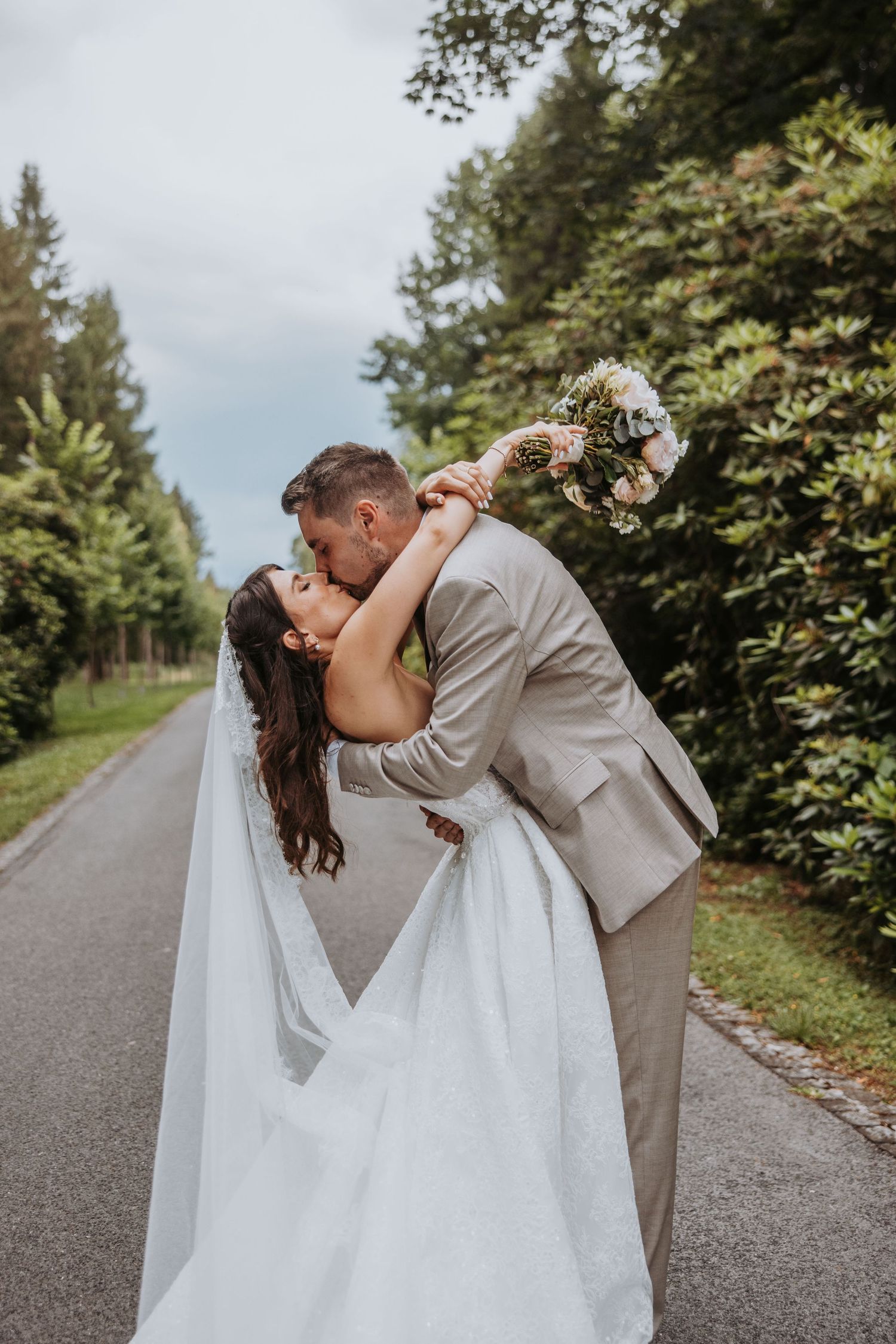 Groom dips bride for romantic kiss on tree-lined road while she holds bridal bouquet.