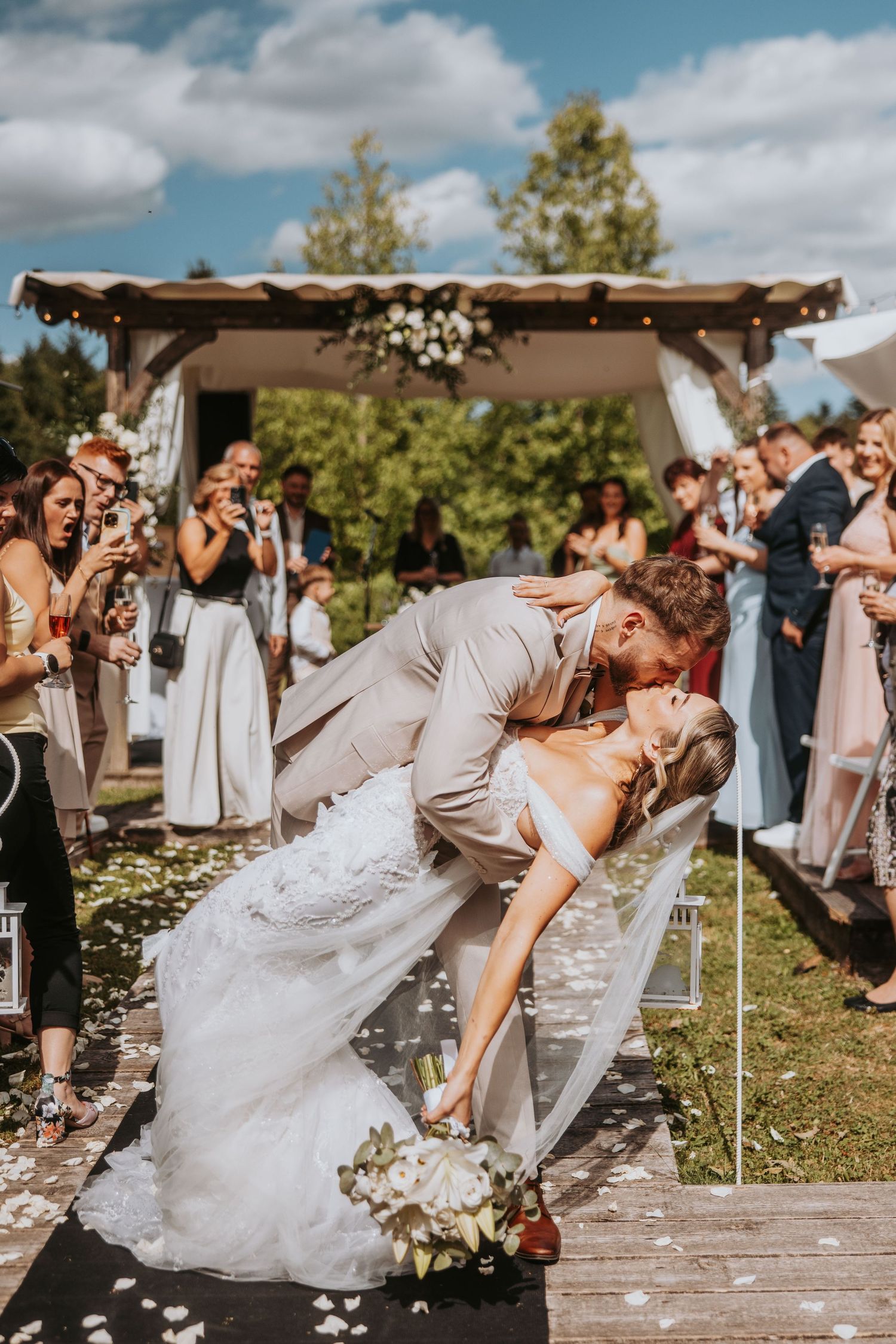 Groom in beige suit dips bride in lace gown for a romantic kiss after outdoor wedding ceremony.