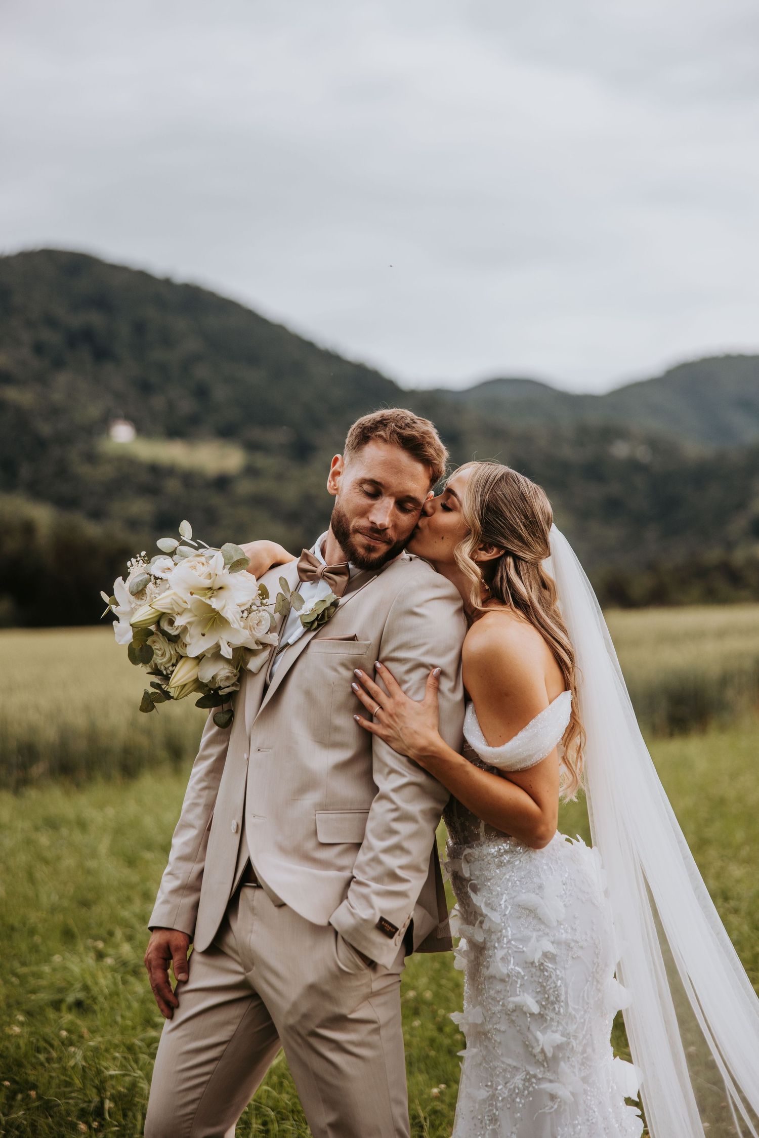 Bride kissing groom on cheek in mountain meadow, groom holding white floral bouquet.