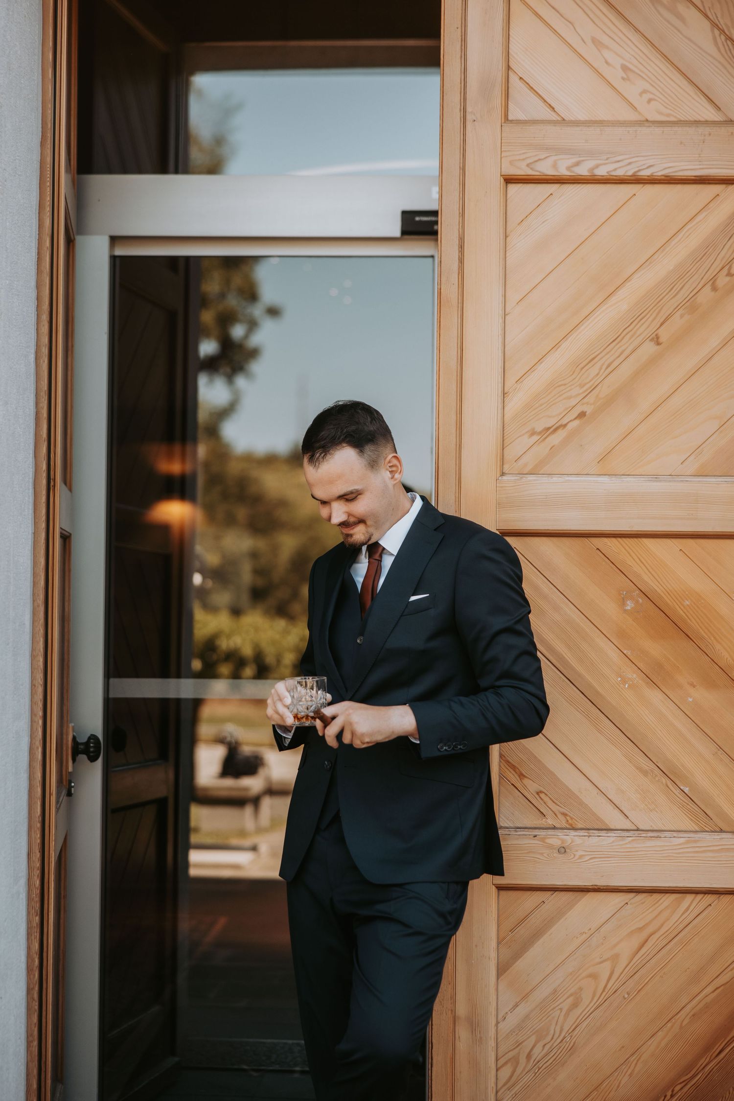 Elegant groom in dark navy suit with burgundy tie smiling while holding cufflinks near wooden door.