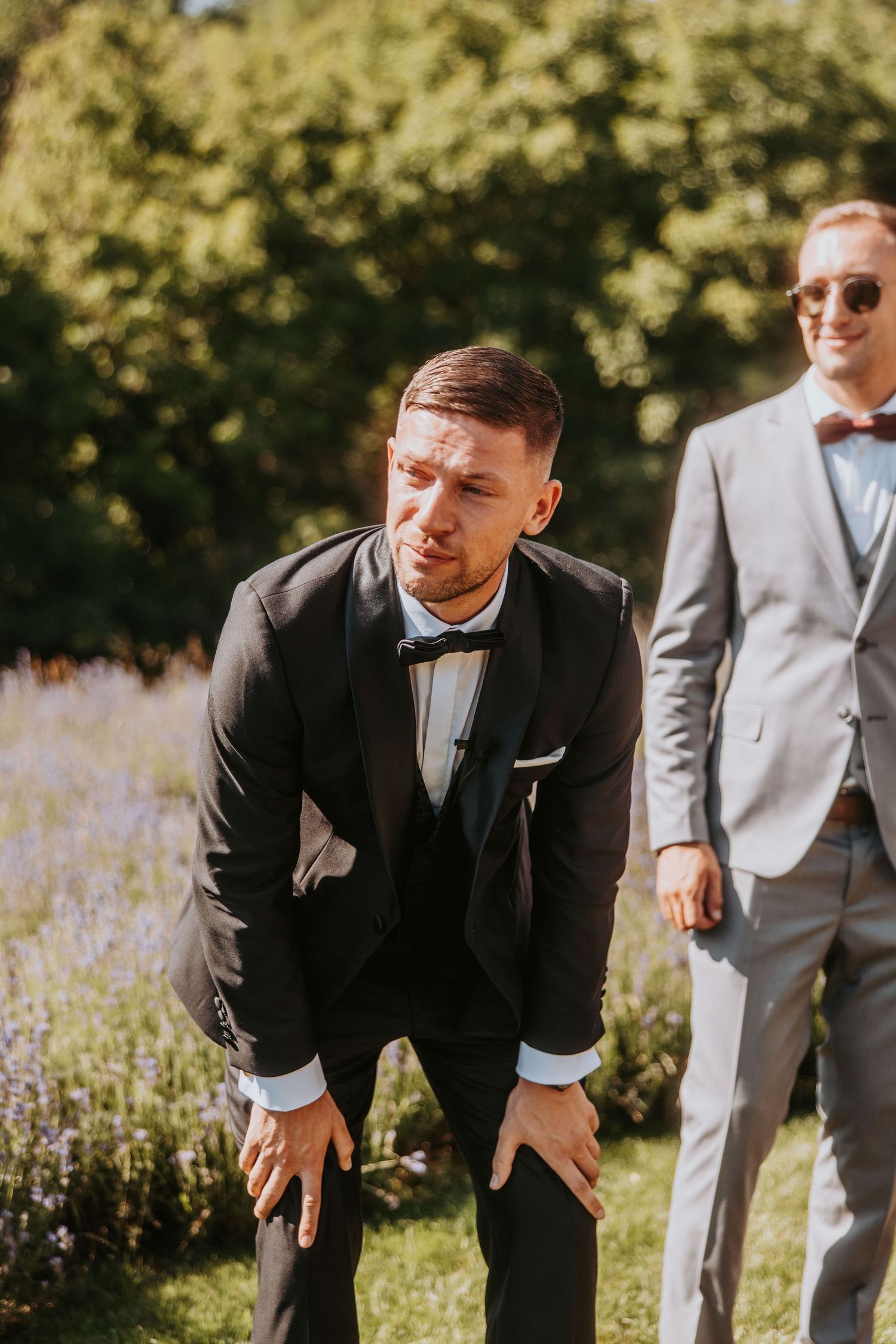 Groom in black tuxedo bowing outdoors with groomsman in gray suit standing behind him at a garden wedding.