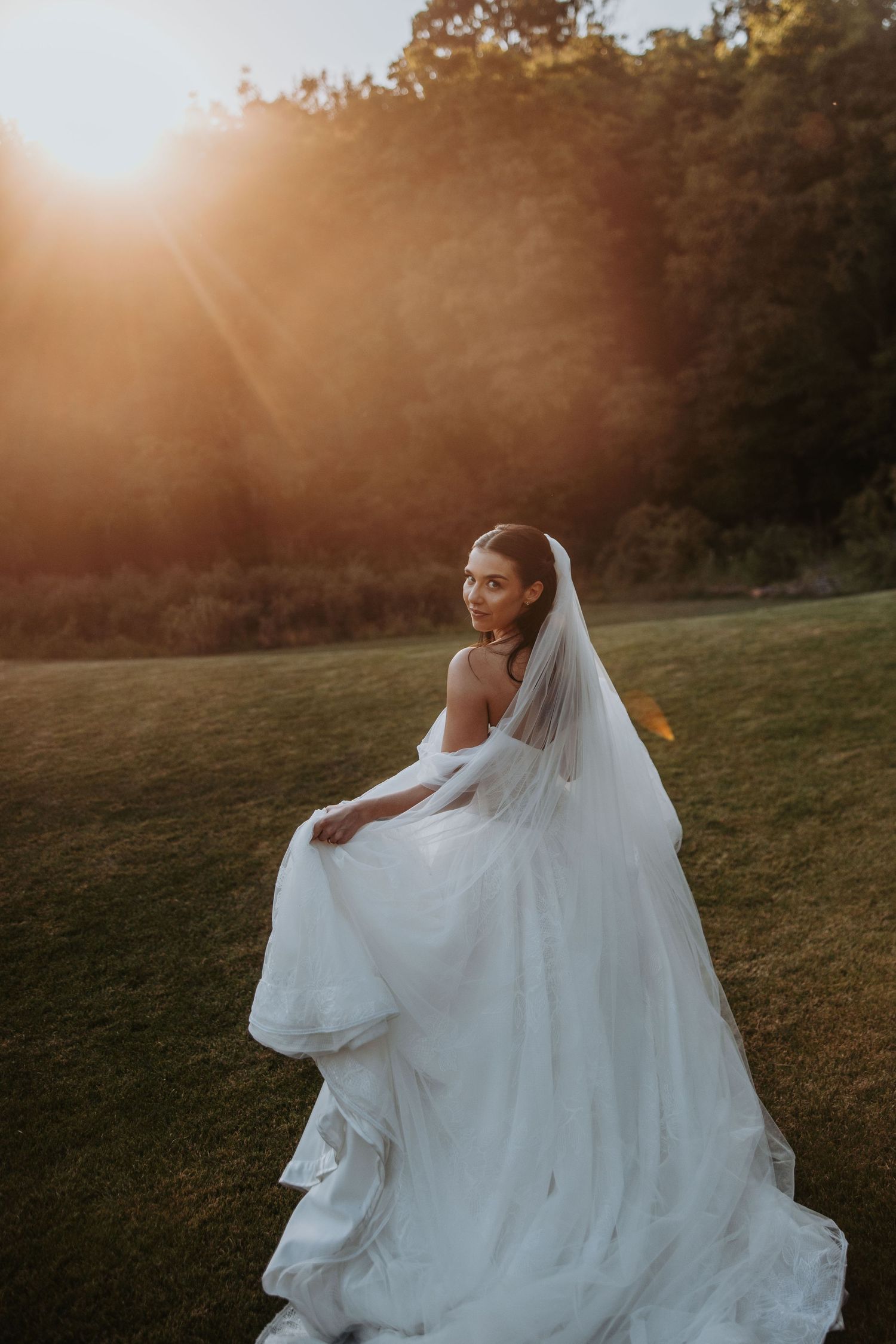Elegant bride in off-shoulder white gown and veil twirling in golden sunset light on green lawn.