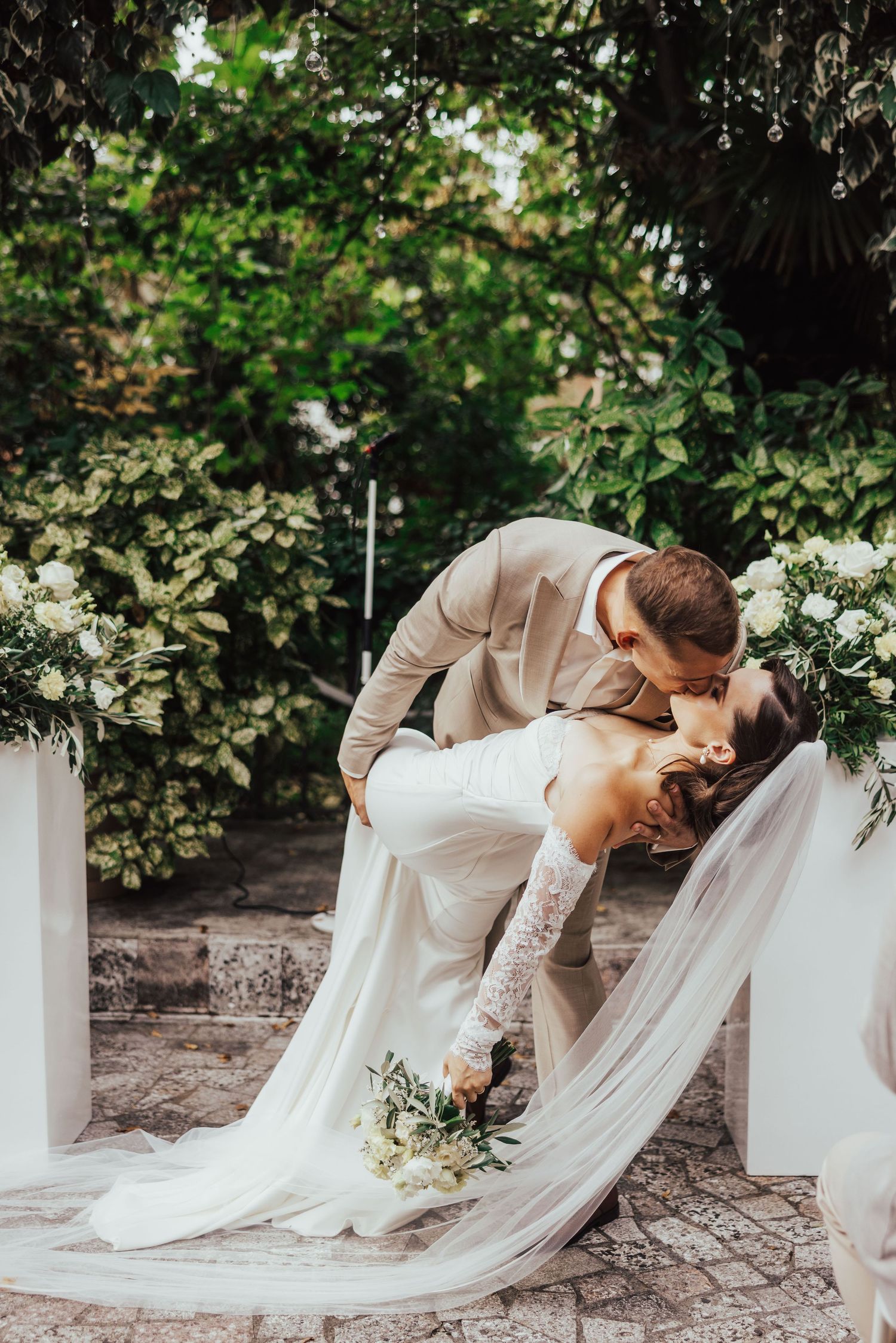 Groom dips bride for romantic kiss at outdoor wedding ceremony surrounded by white floral arrangements.