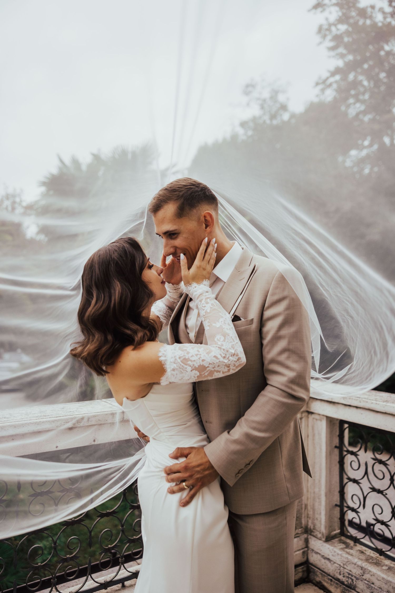 Bride and groom sharing a romantic kiss under a flowing veil on a balcony, bride in lace gown, groom in beige suit.