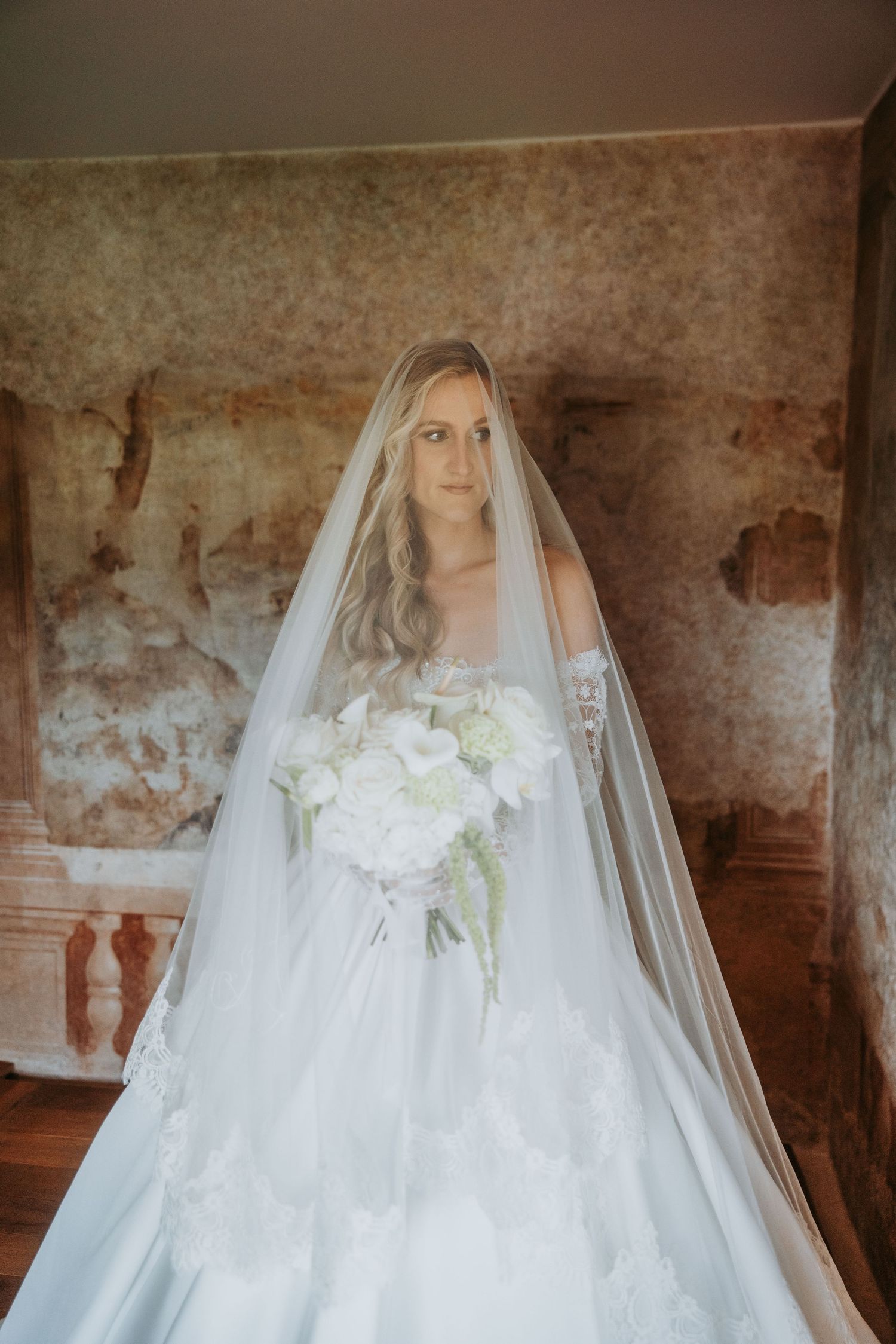 Elegant bride in strapless lace gown with long veil holding white floral bouquet indoors.