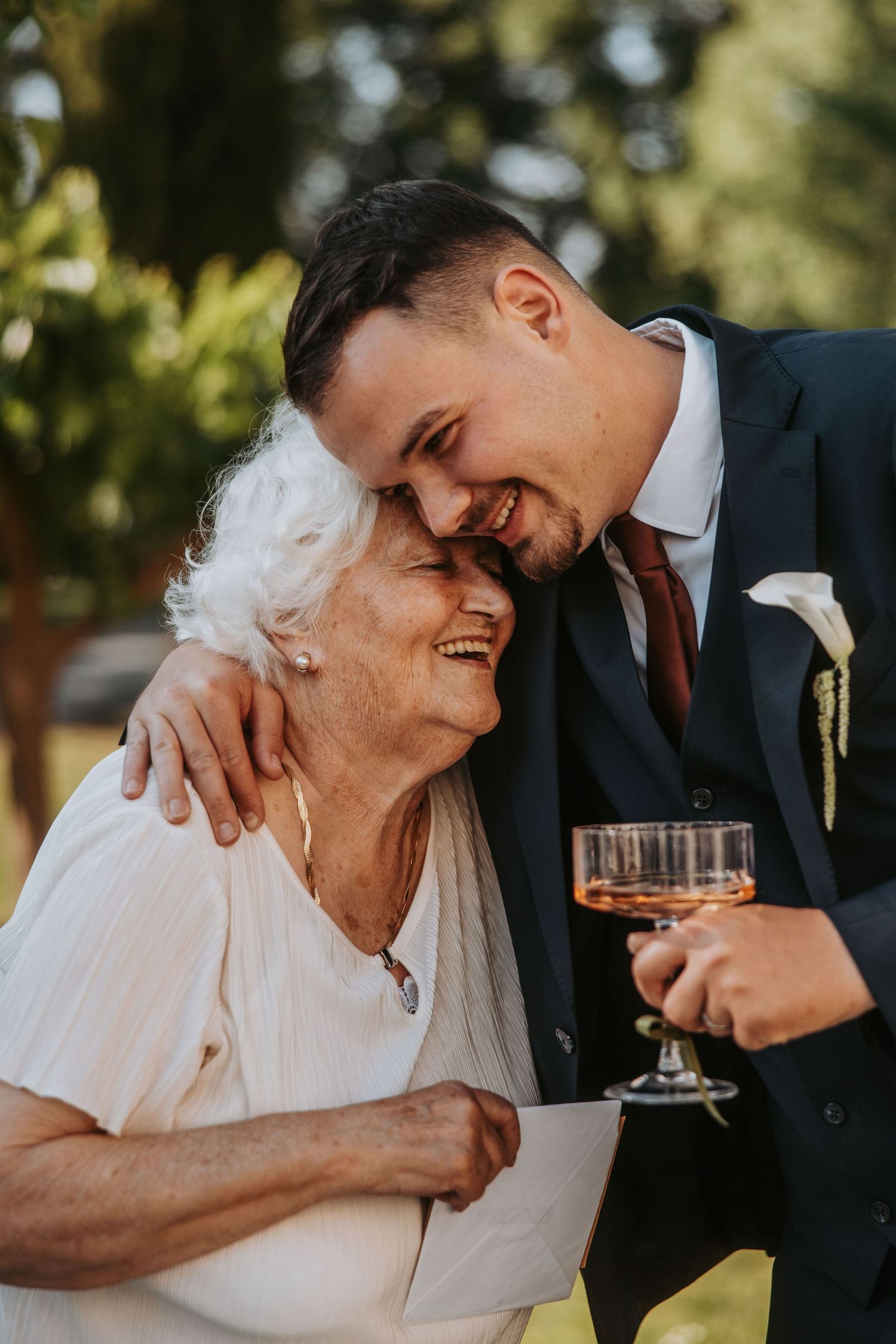 Groom in navy suit embracing elderly grandmother while holding a rose gold champagne glass outdoors.