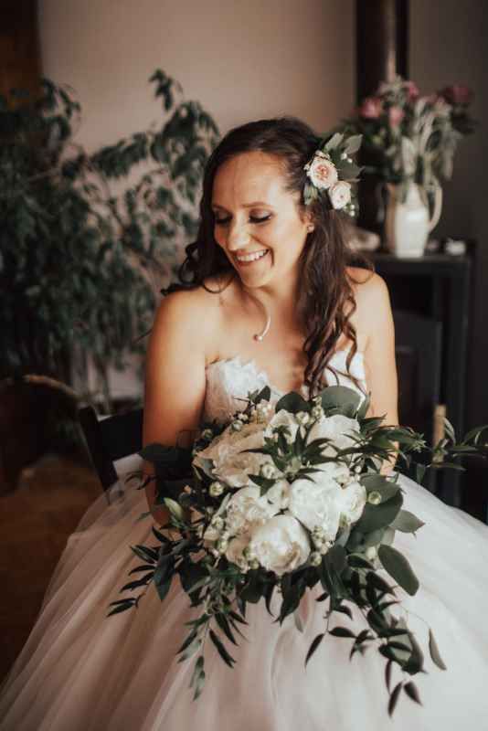 Smiling brunette bride in strapless ballgown holds white peony bouquet with greenery indoors.