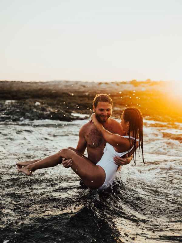 Happy couple playing in ocean waves at sunset, man lifting woman in white swimsuit.