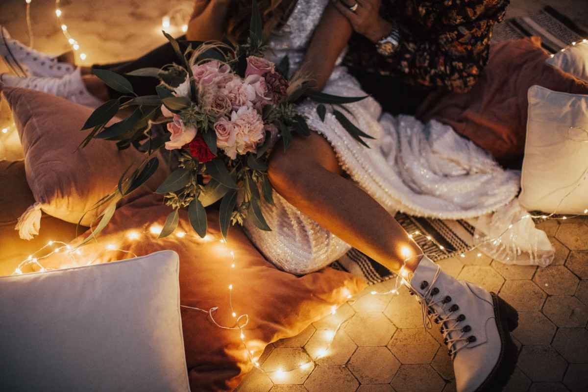 A bride sits surrounded by fairy lights holding a pink floral bouquet wearing white lace dress and boots.