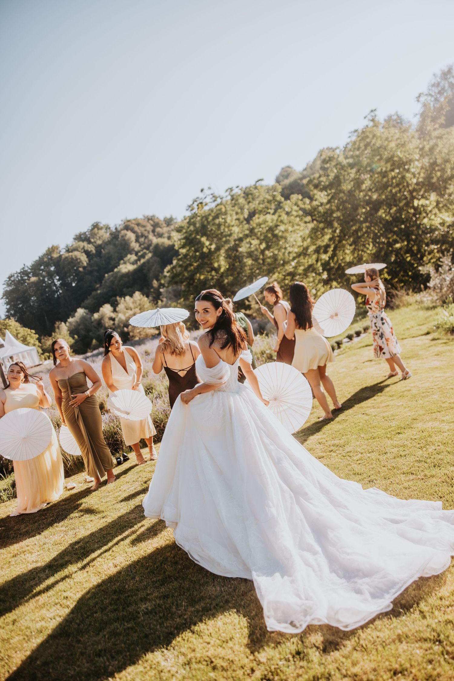 Bride in a flowing white gown twirls on a sunny lawn surrounded by guests holding decorative parasols.