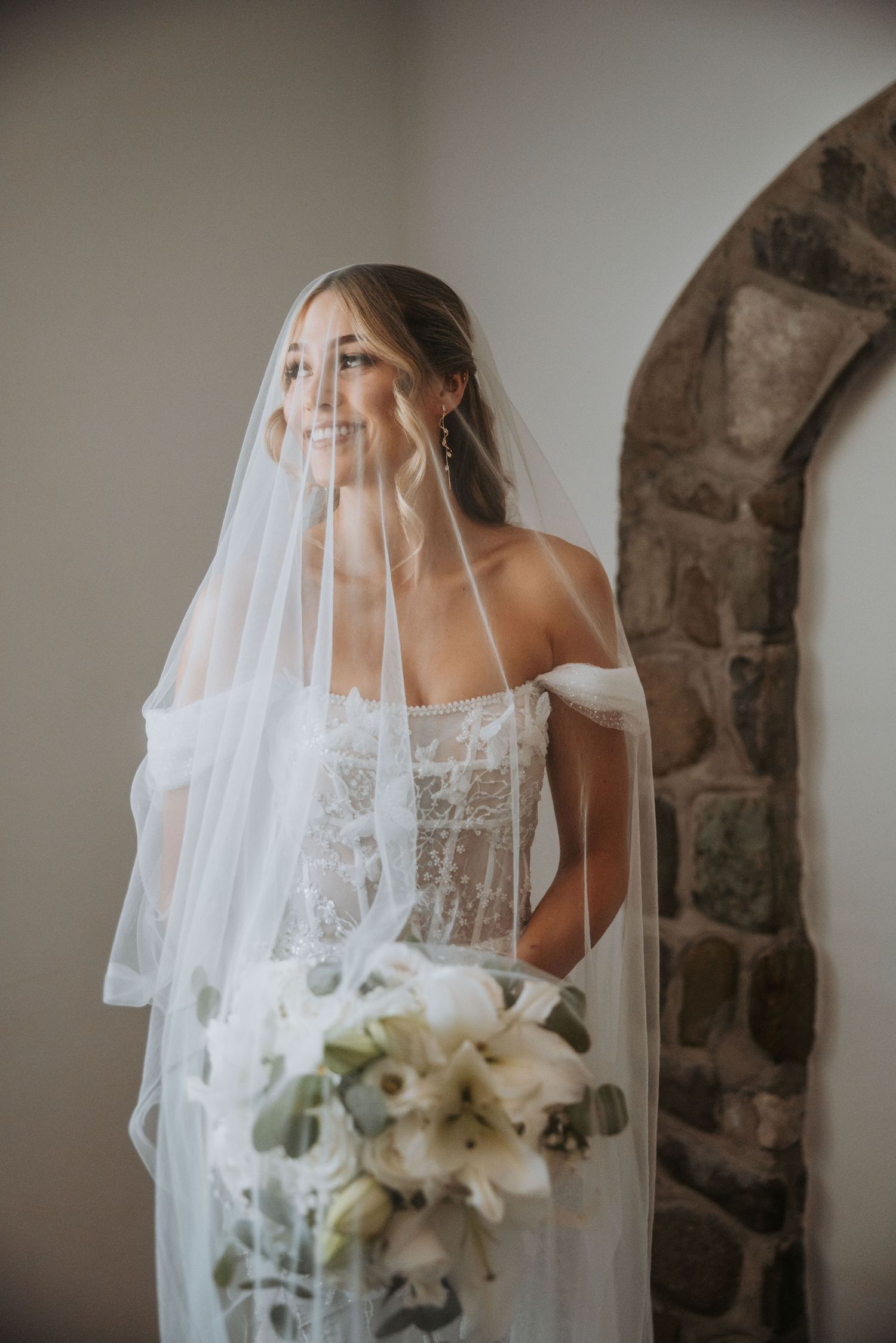 Smiling bride in off-shoulder lace gown holding white floral bouquet with flowing veil draped over her.