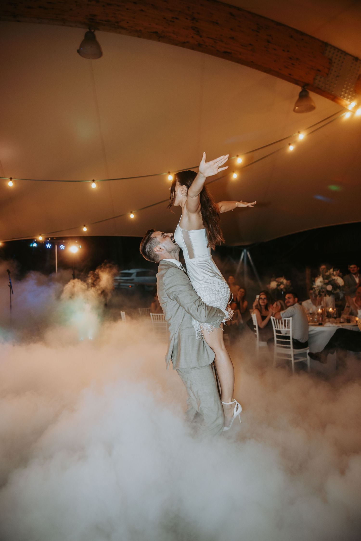 Groom lifting bride dramatically during first dance surrounded by fog and string lights.