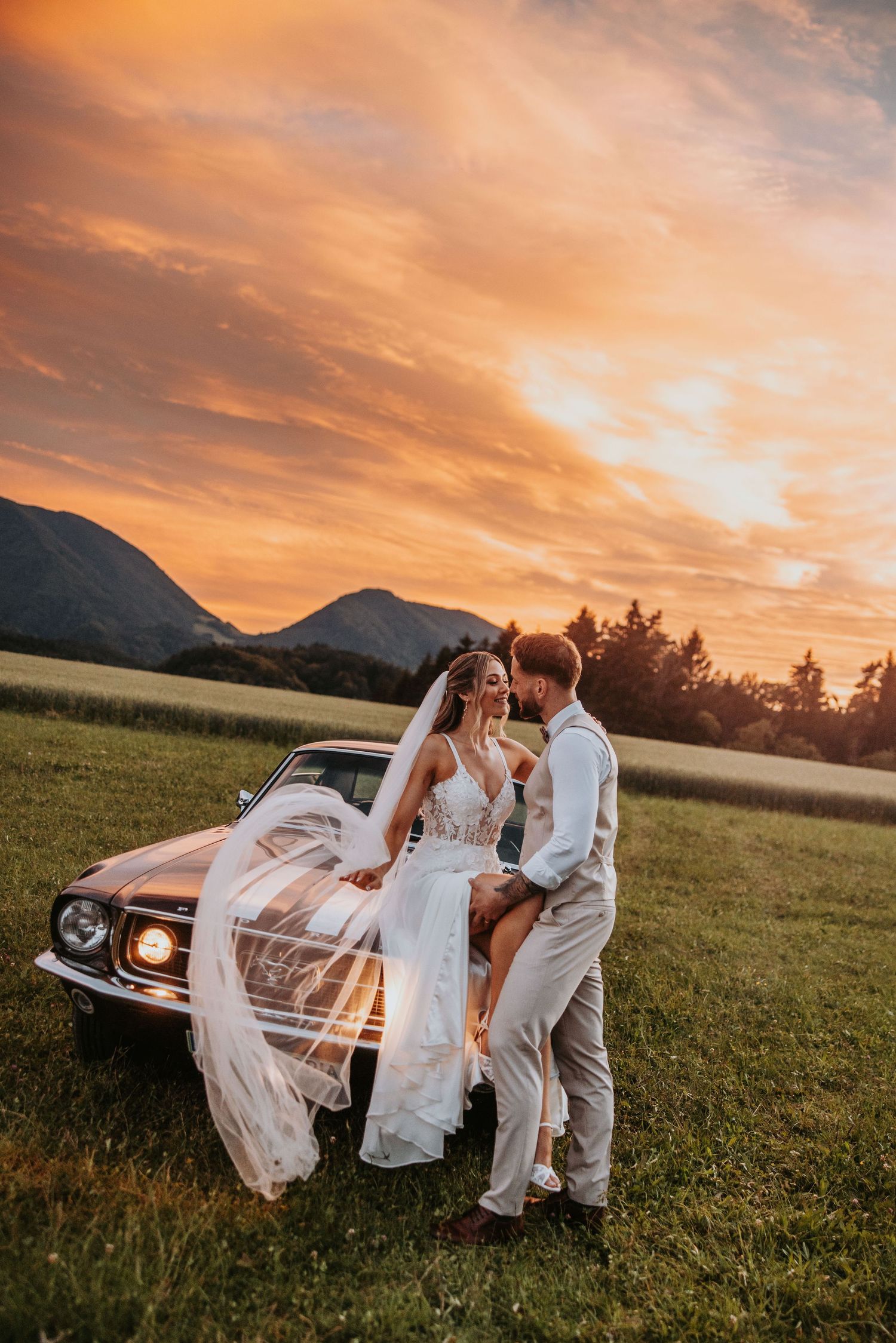 Groom lifts bride onto vintage car hood as her veil flows in wind at dramatic golden sunset.