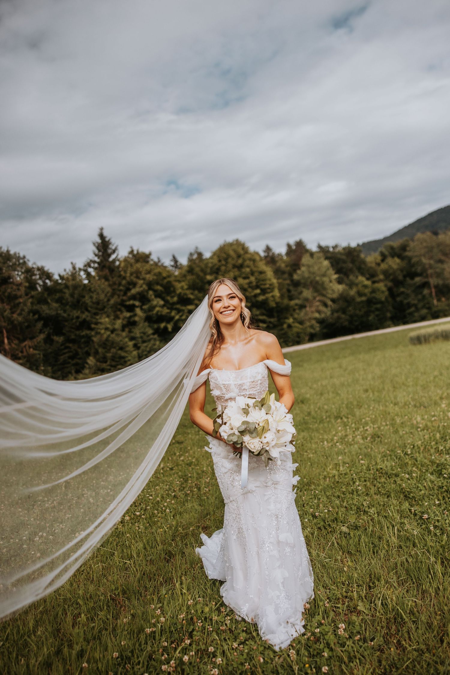 Smiling bride in lace dress with long veil blowing in wind in scenic meadow.