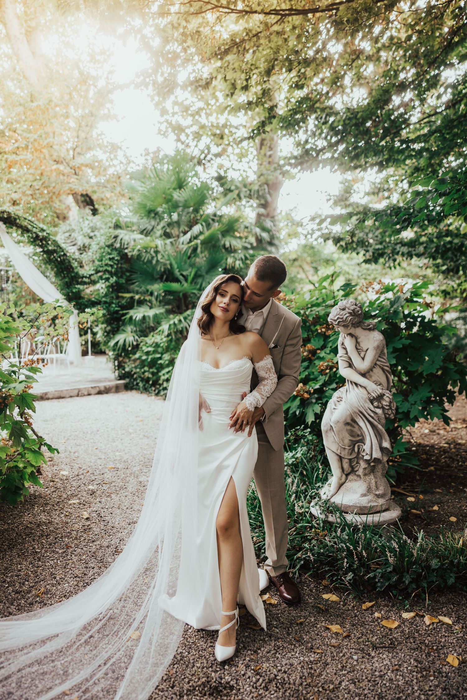 Bride in fitted white gown with long veil stands with groom in sunlit garden near stone statue.