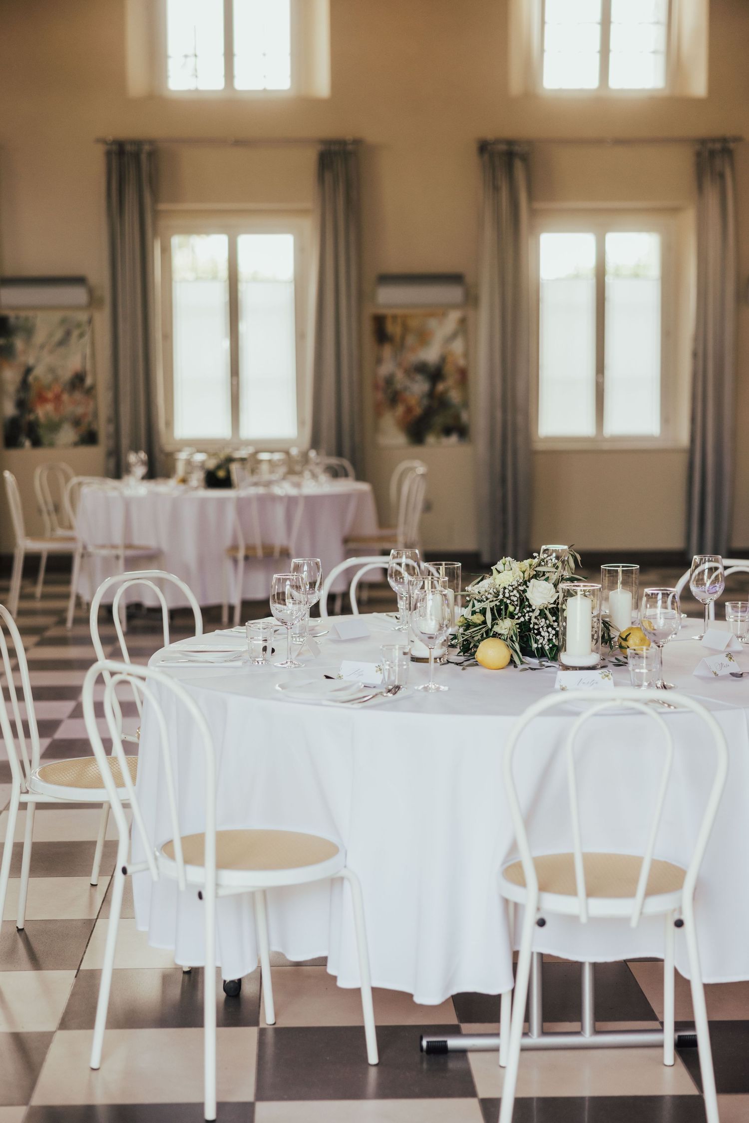 Elegant wedding reception hall with white round tables, Thonet chairs, floral centerpieces, and tall windows with soft natural light.