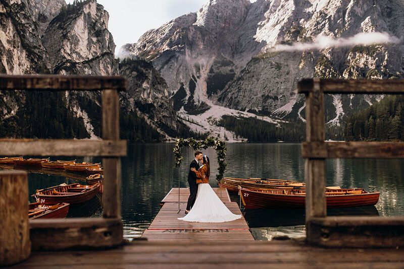 Wedding couple kissing on a wooden dock at Lago di Braies surrounded by mountains and red boats.