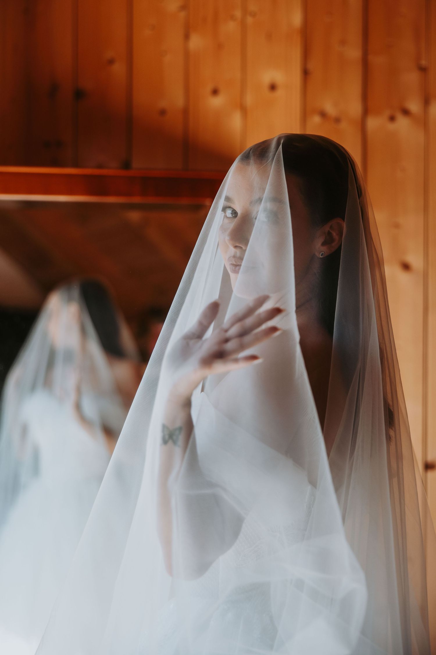 A beautiful bride draped in a sheer white veil gazing forward with a butterfly tattoo visible on her hand.