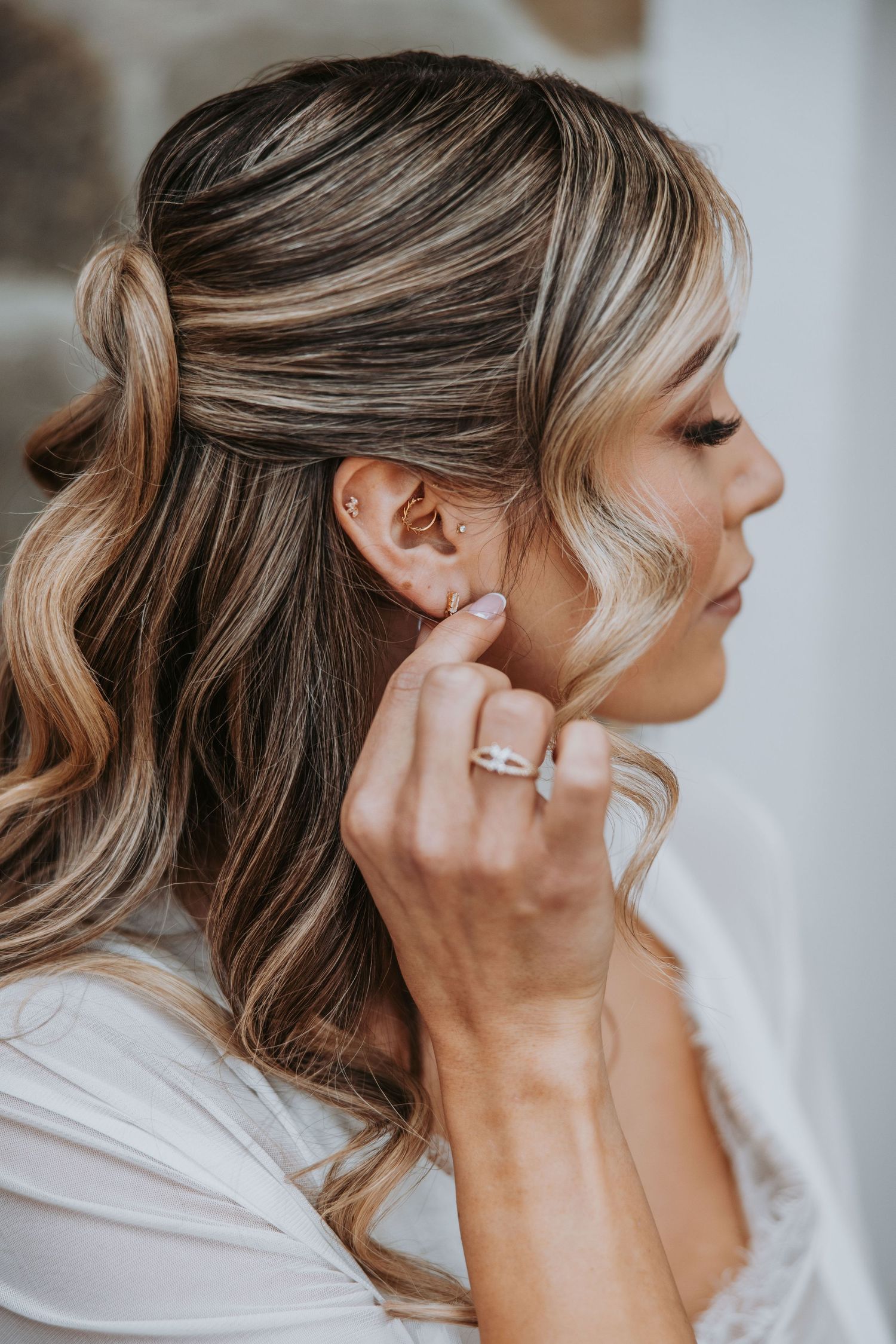 Bride with highlighted hair adjusts earring wearing engagement ring during wedding day preparations.