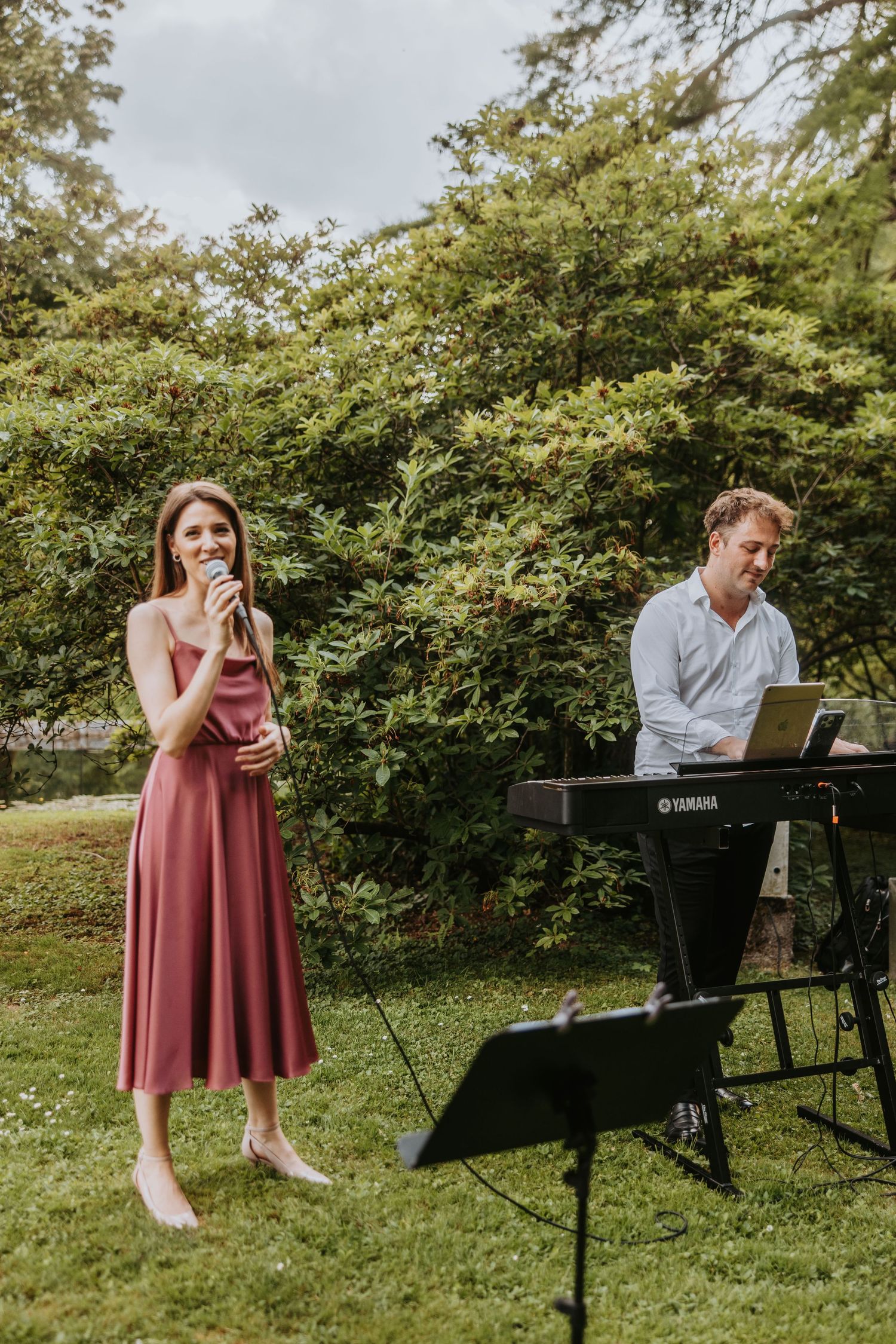 Female singer in pink dress performs outdoors with male pianist at Yamaha keyboard.
