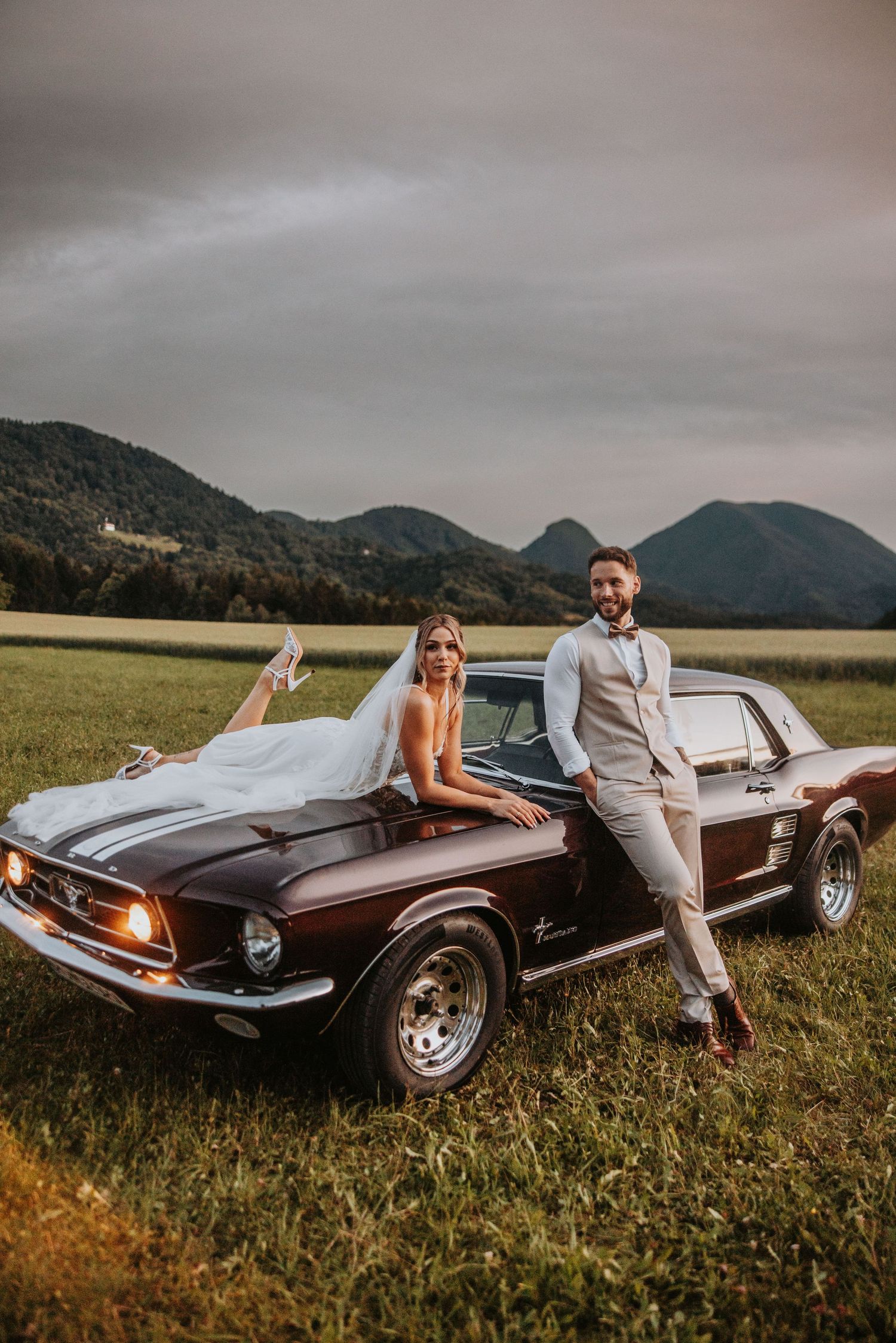 Bride and groom posing with a vintage dark red Ford Mustang in a mountain field at dusk.