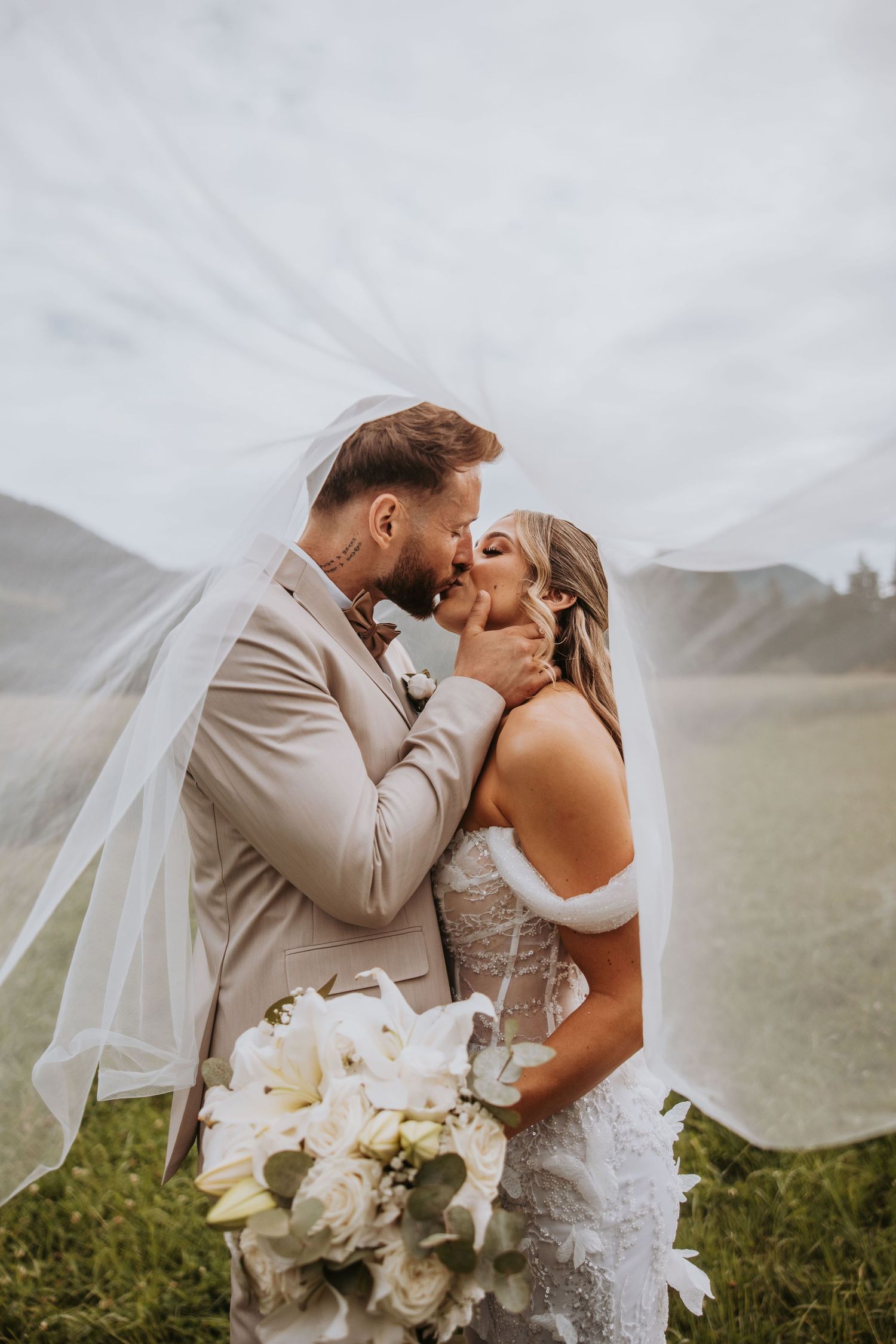 Bride and groom share a tender kiss under flowing veil with white floral bouquet in scenic outdoor setting.