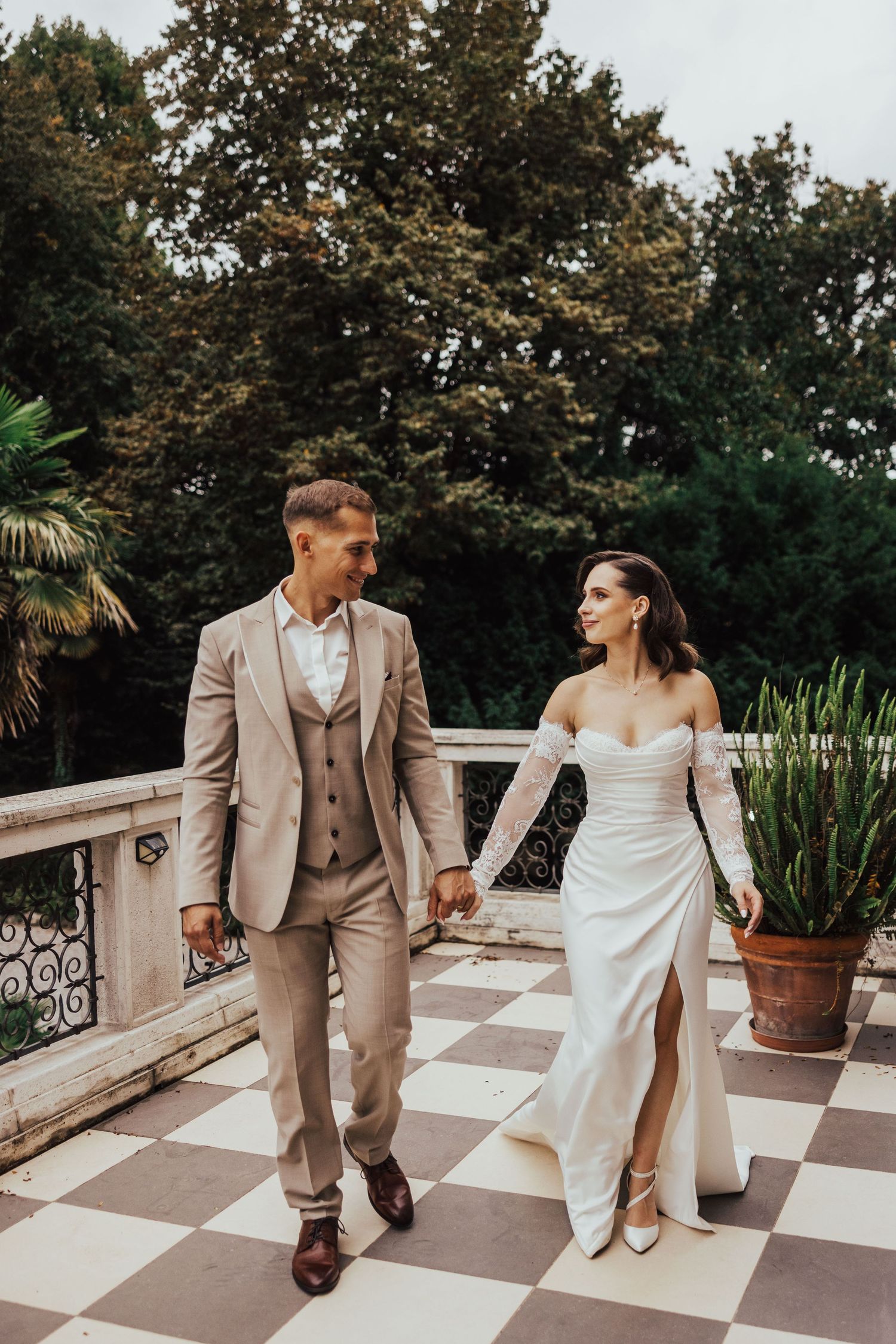 Bride and groom holding hands walking on checkered terrace with lush trees.
