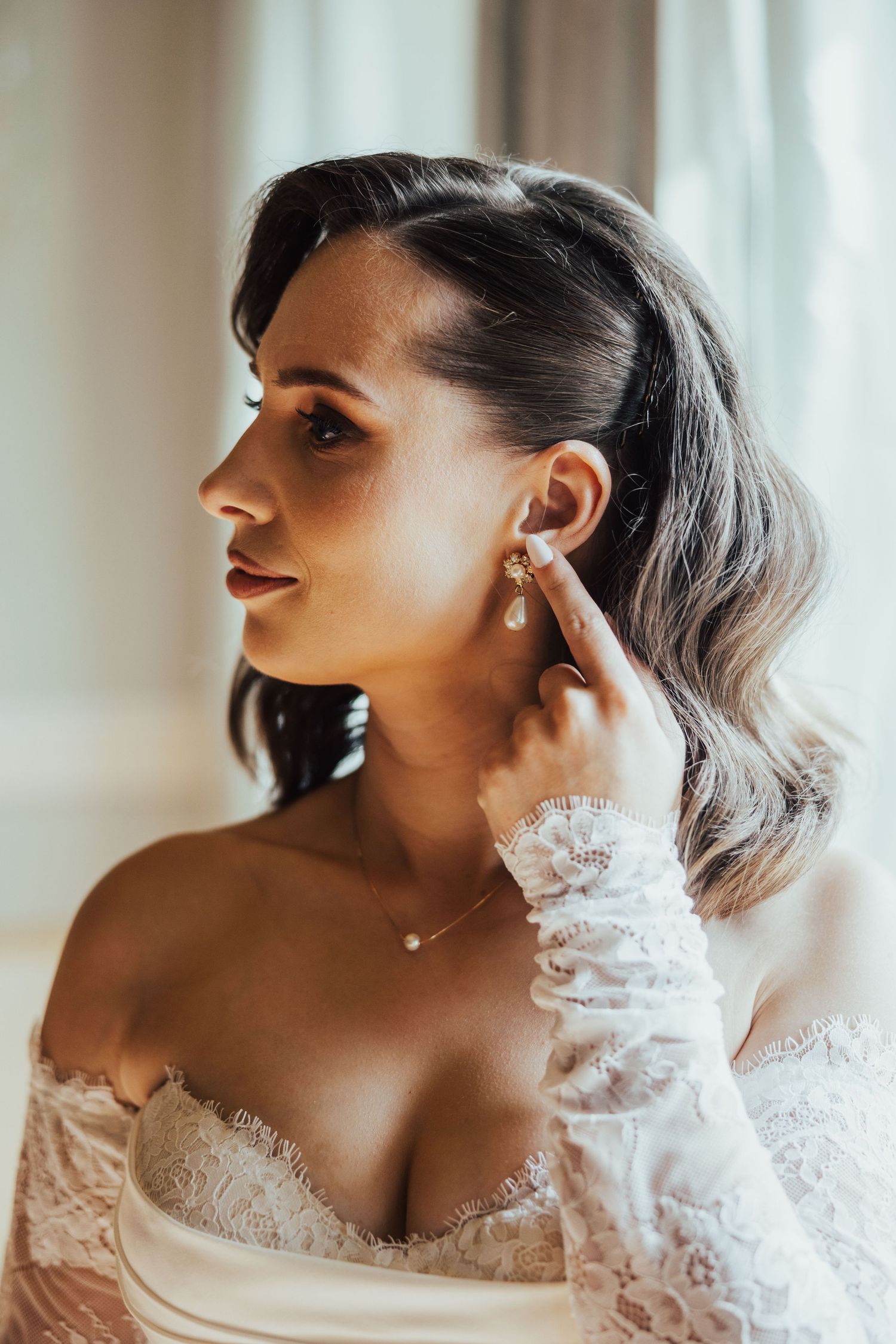 Bride adjusting pearl drop earring in lace off-shoulder gown, soft natural light, elegant bridal jewelry and necklace.