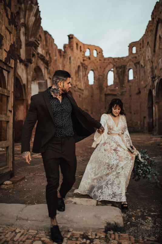 Couple in wedding attire walking through ancient stone ruins, bride in lace gown holding bouquet, groom in black suit.