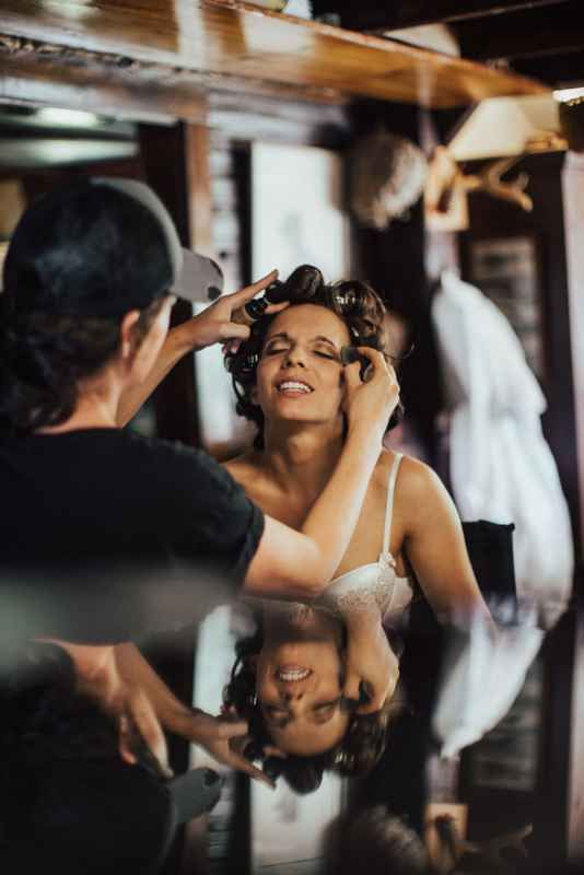 Bride smiling in mirror reflection while hairstylist sets her hair in curlers during wedding preparation.