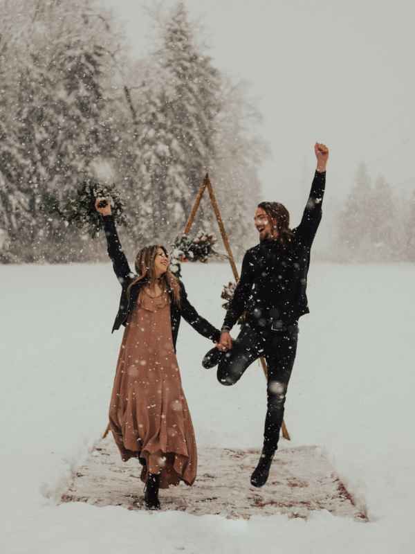 Joyful couple jumping and celebrating in the snow during a winter wedding shoot with snowy trees behind.