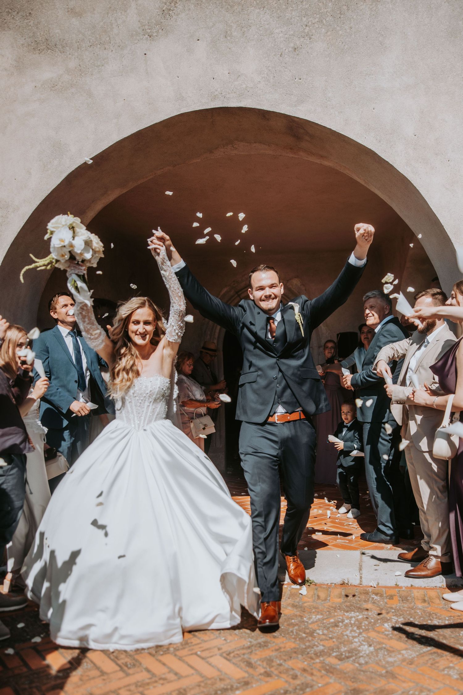Joyful newlyweds exit under stone arch as guests toss white flower petals at outdoor wedding.