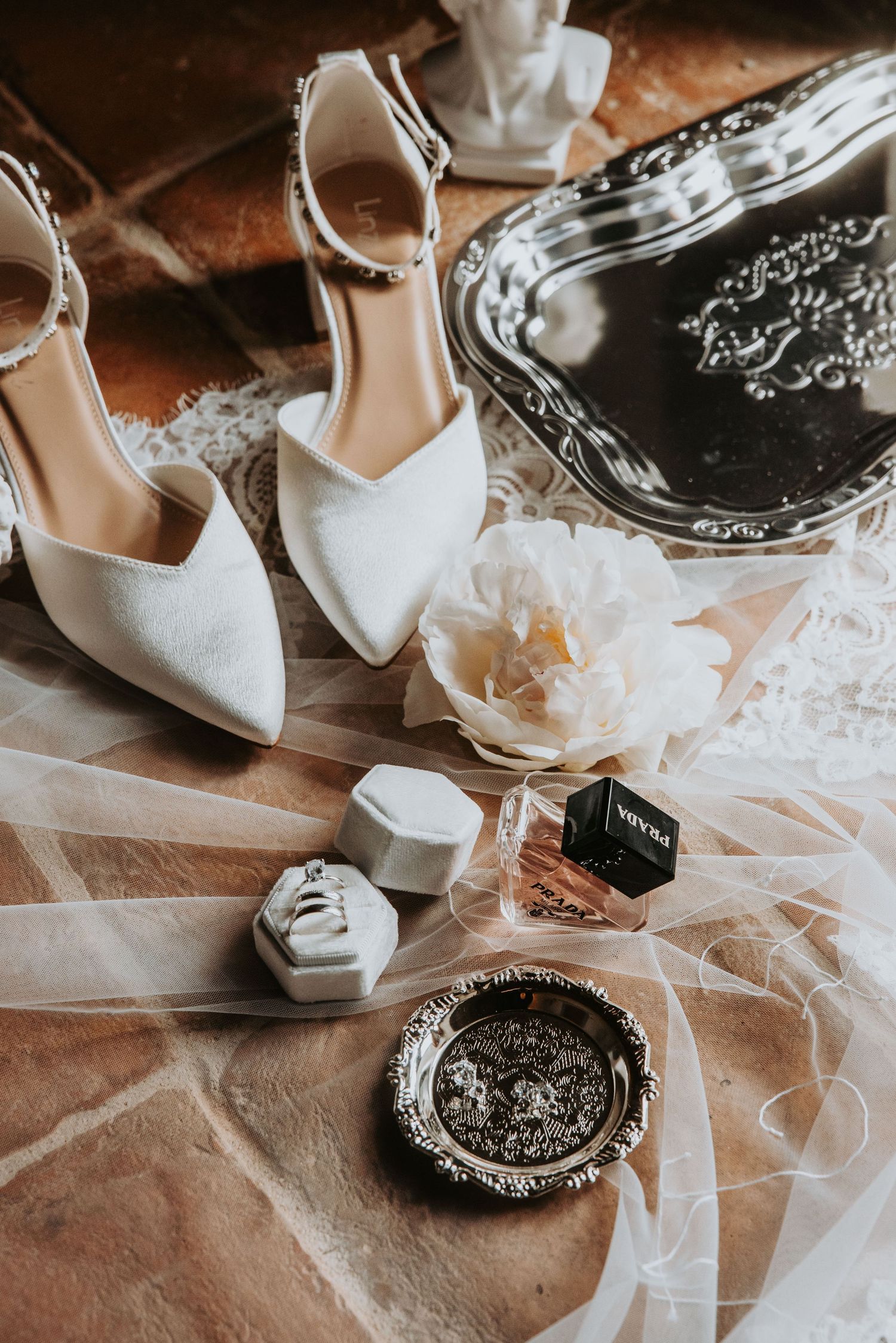 Elegant bridal accessories flat lay with white heels, ring box, perfume, and veil on terracotta floor.