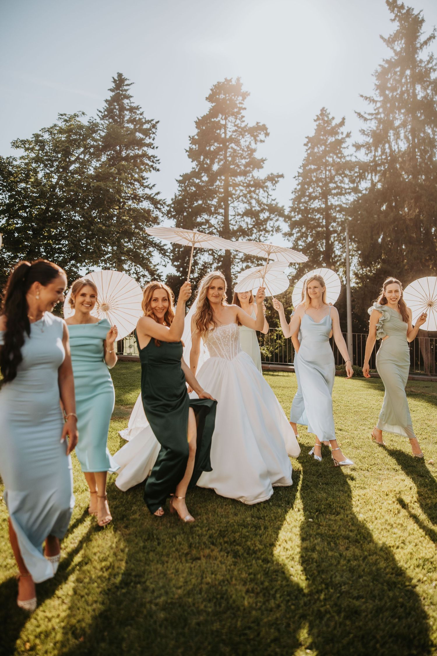 Bride and bridesmaids in blue dresses walking on grass holding white parasols in sunlight.