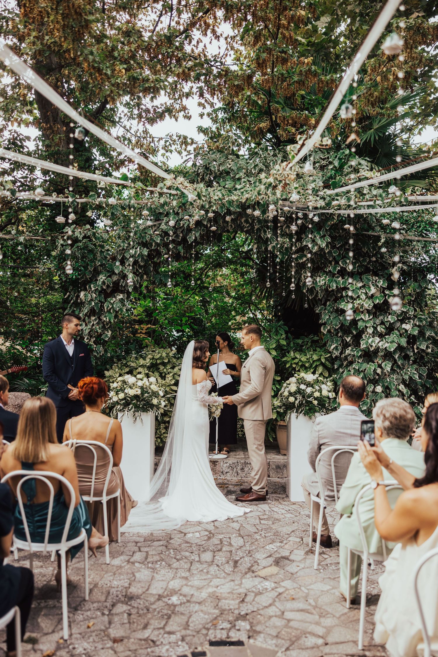 Bride and groom exchanging vows under floral arch surrounded by wedding guests outdoors.