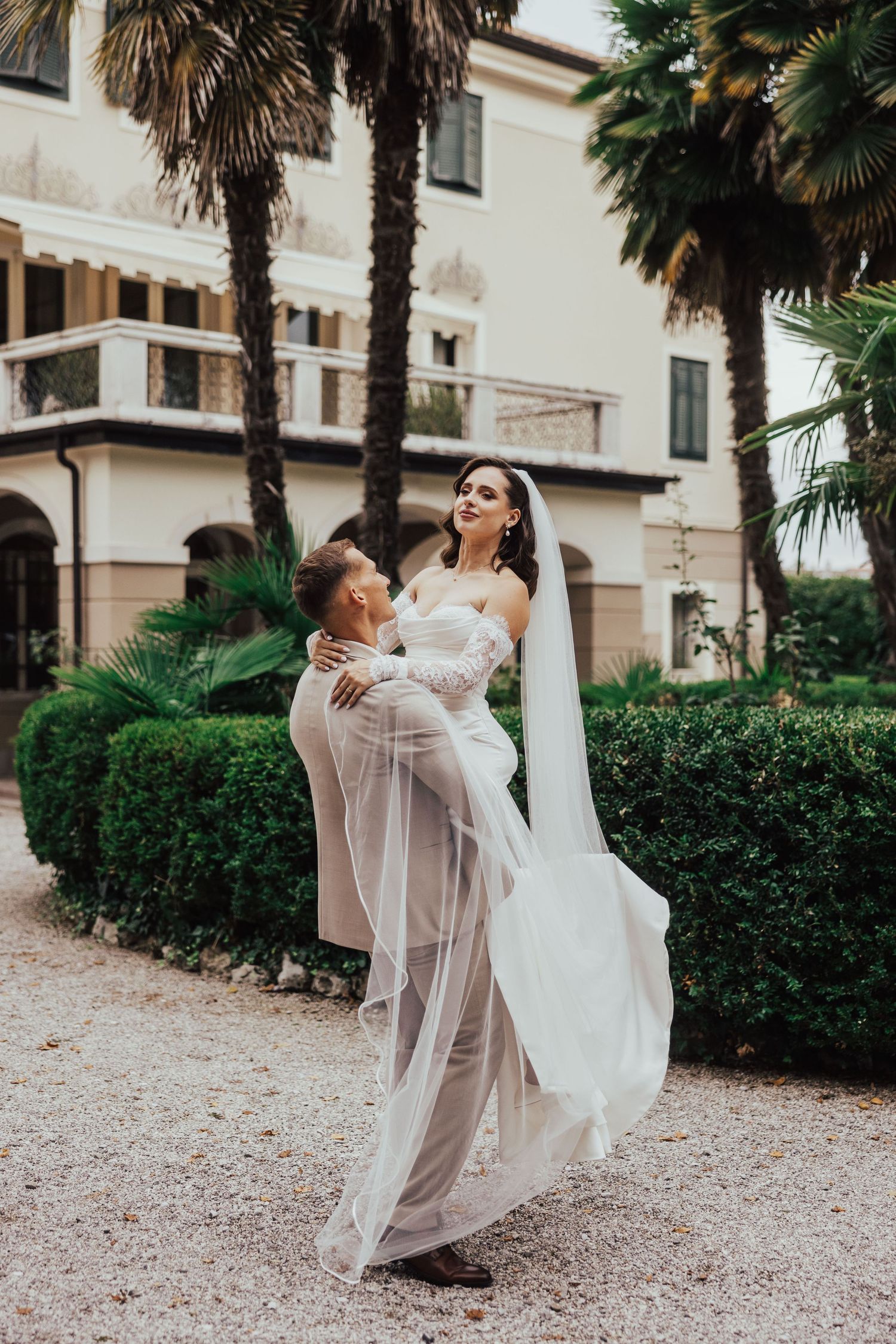 Groom lifting bride in white wedding dress and veil outdoors near palm trees and elegant villa.